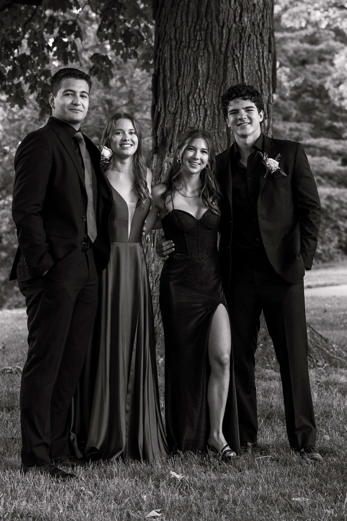 Four formally dressed young adults posing together near a tree outdoors in a wedding group portrait