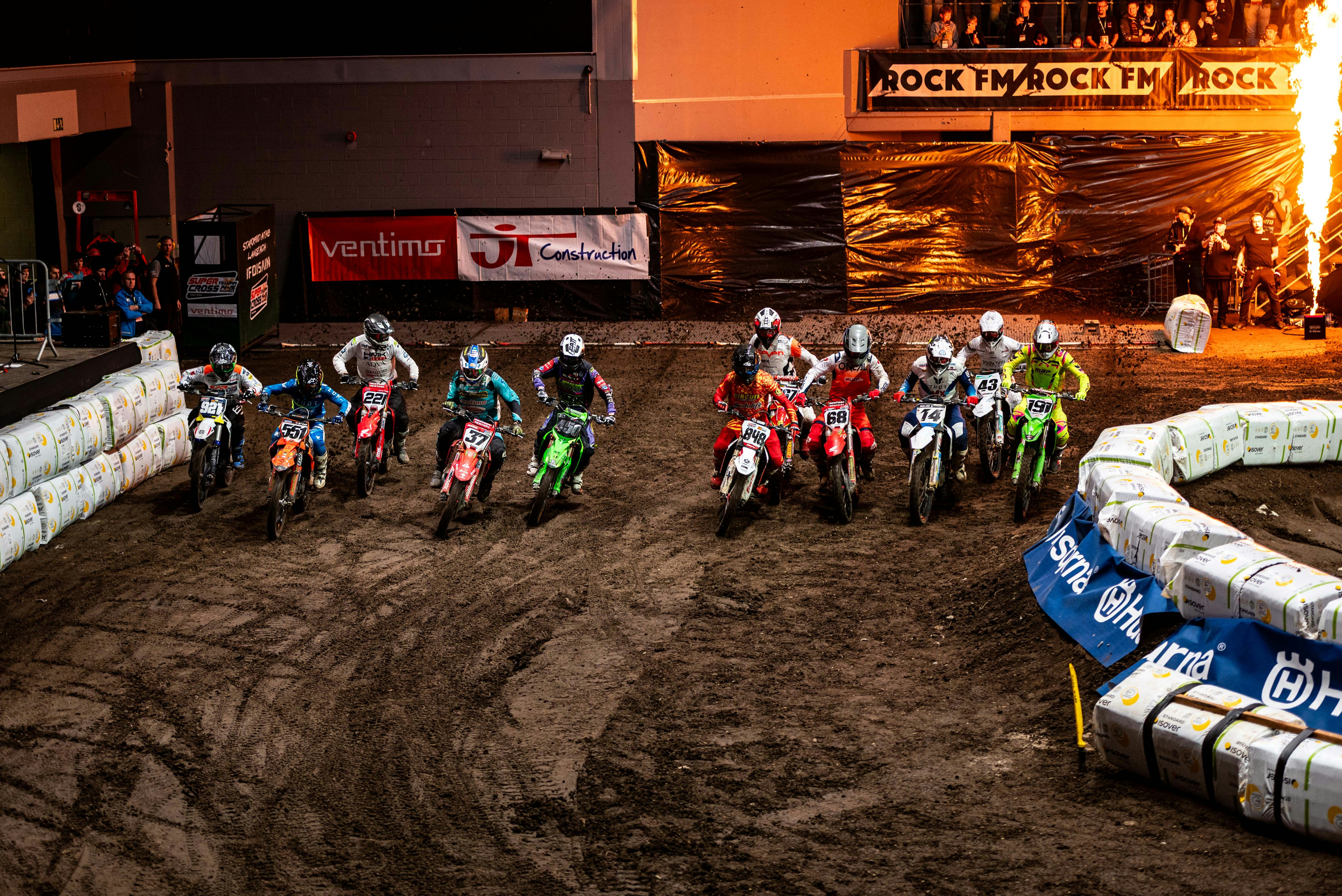 Motocross riders race on a dirt track at night.