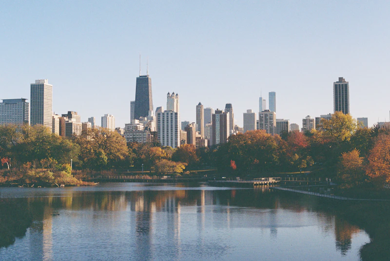 Autumn foliage around a lagoon with the Chicago skyline, including the John Hancock Center, reflected in the water