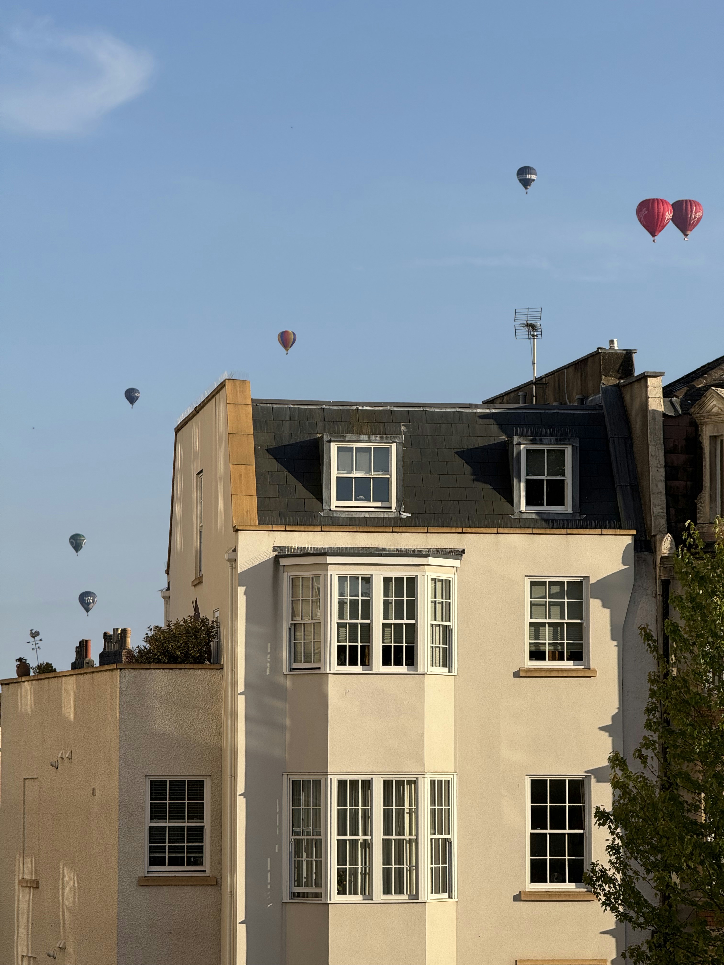Hot air balloons float over buildings under a clear sky.