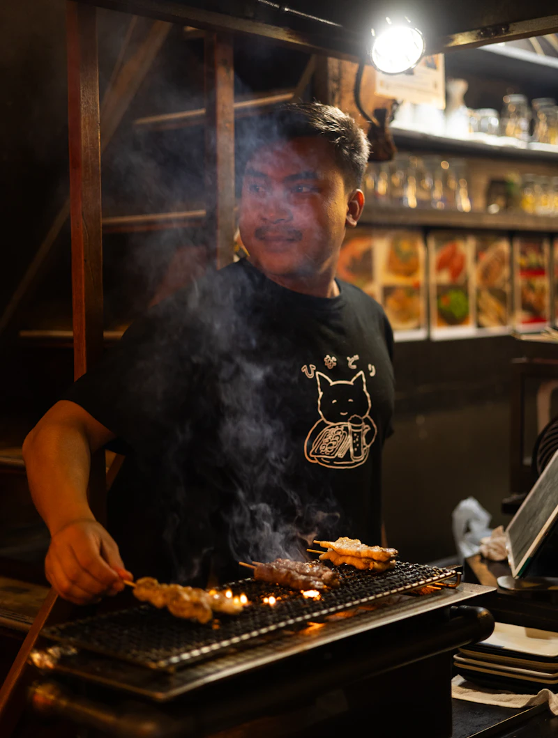 A cook turns yakitori skewers over a charcoal grill behind a counter, smoke rising between him and the seats