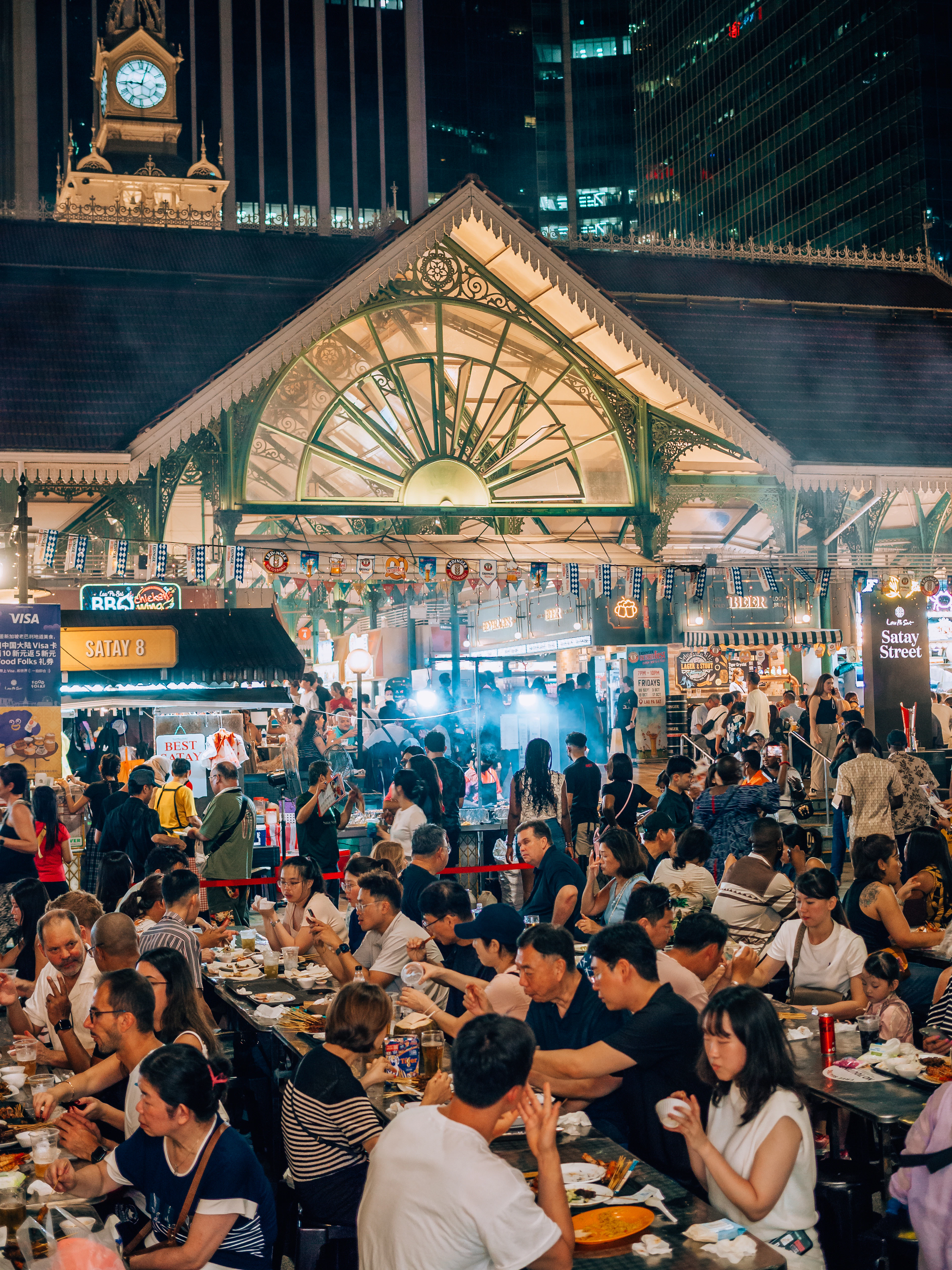 Diners packed around tables on a night-time satay street beside the Victorian cast-iron pavilion of a Singapore hawker centre, skyscrapers lit up behind