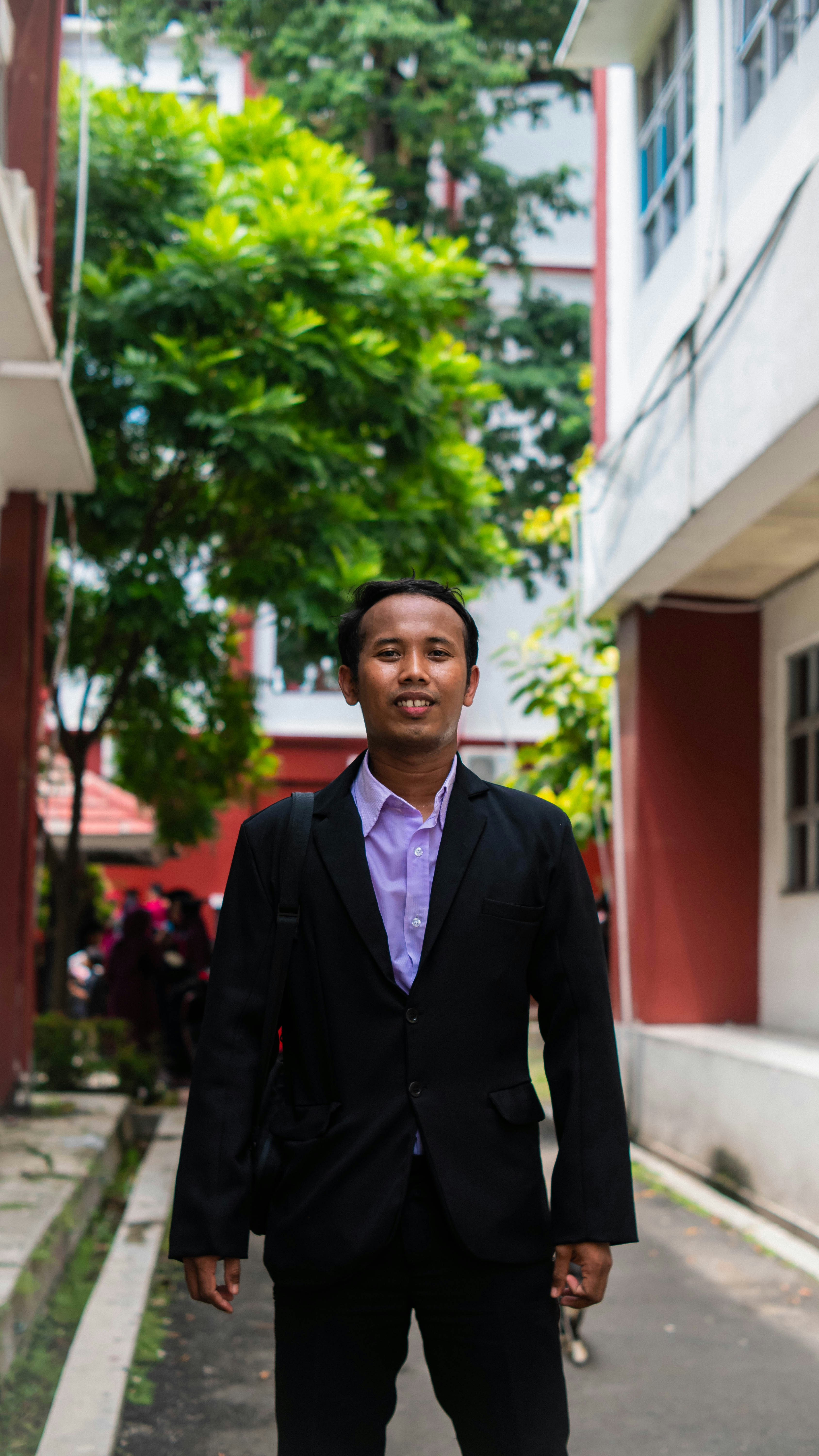 A man in a suit stands between buildings with trees.