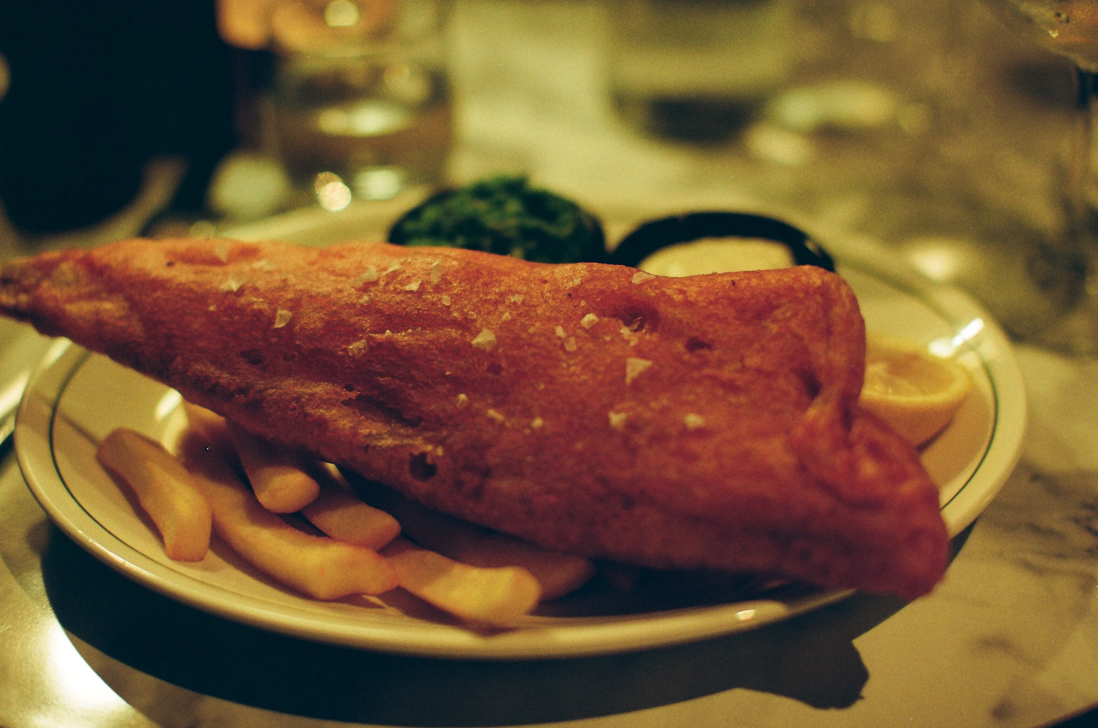 A large piece of batter-fried fish on a plate of chips with mushy peas, tartare sauce and a lemon wedge, photographed in warm chip-shop light