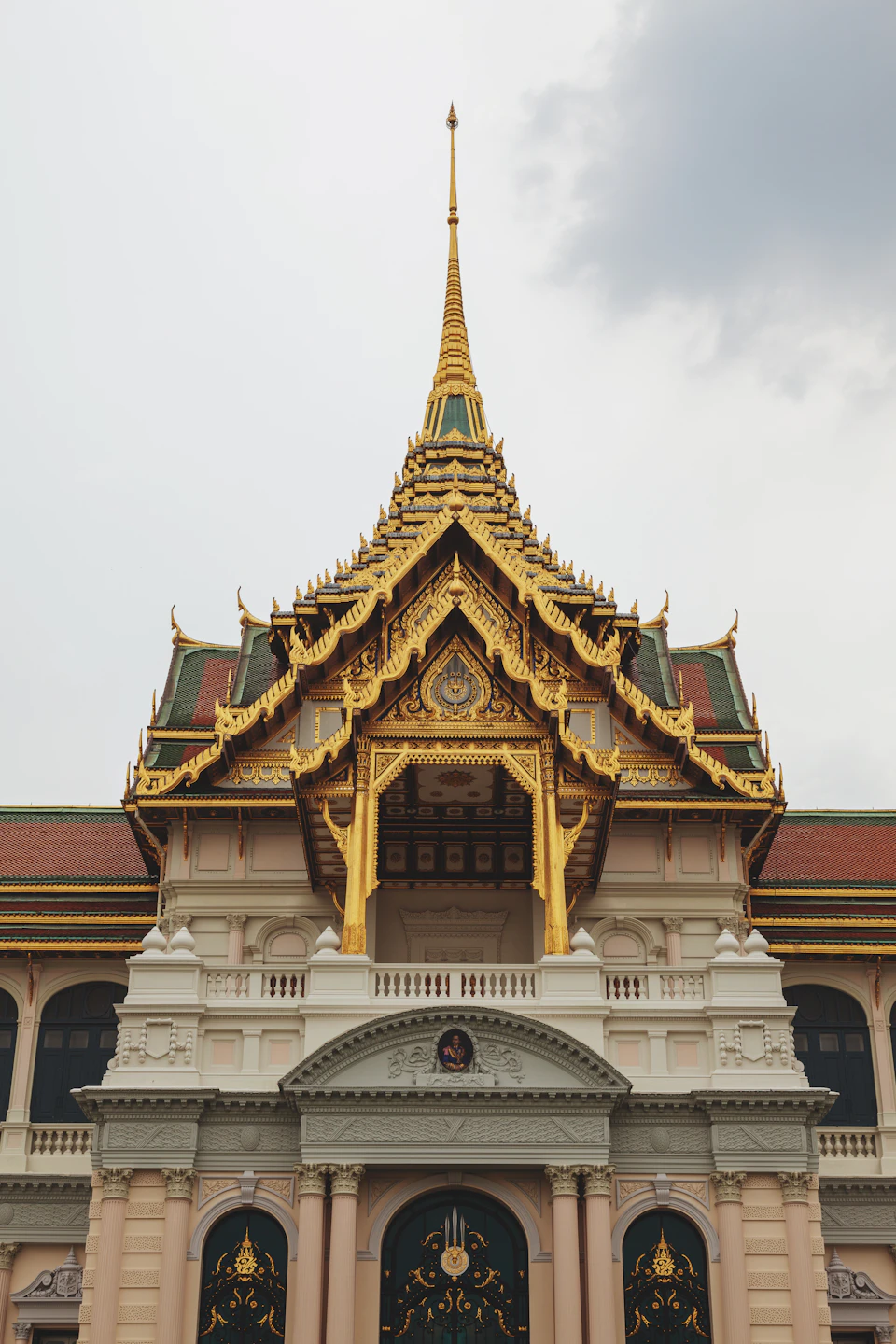 The image shows a majestic temple with a golden roof and intricate decorations, standing tall against an overcast sky. The temple is adorned with multiple spires and columns, showcasing its grandeur and architectural prowess. The perspective of the photo is from below, looking up at the temple's towering presence.