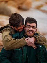 Two smiling men embracing outdoors with rocky background.