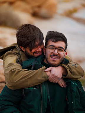 Two smiling men embracing outdoors with rocky background.