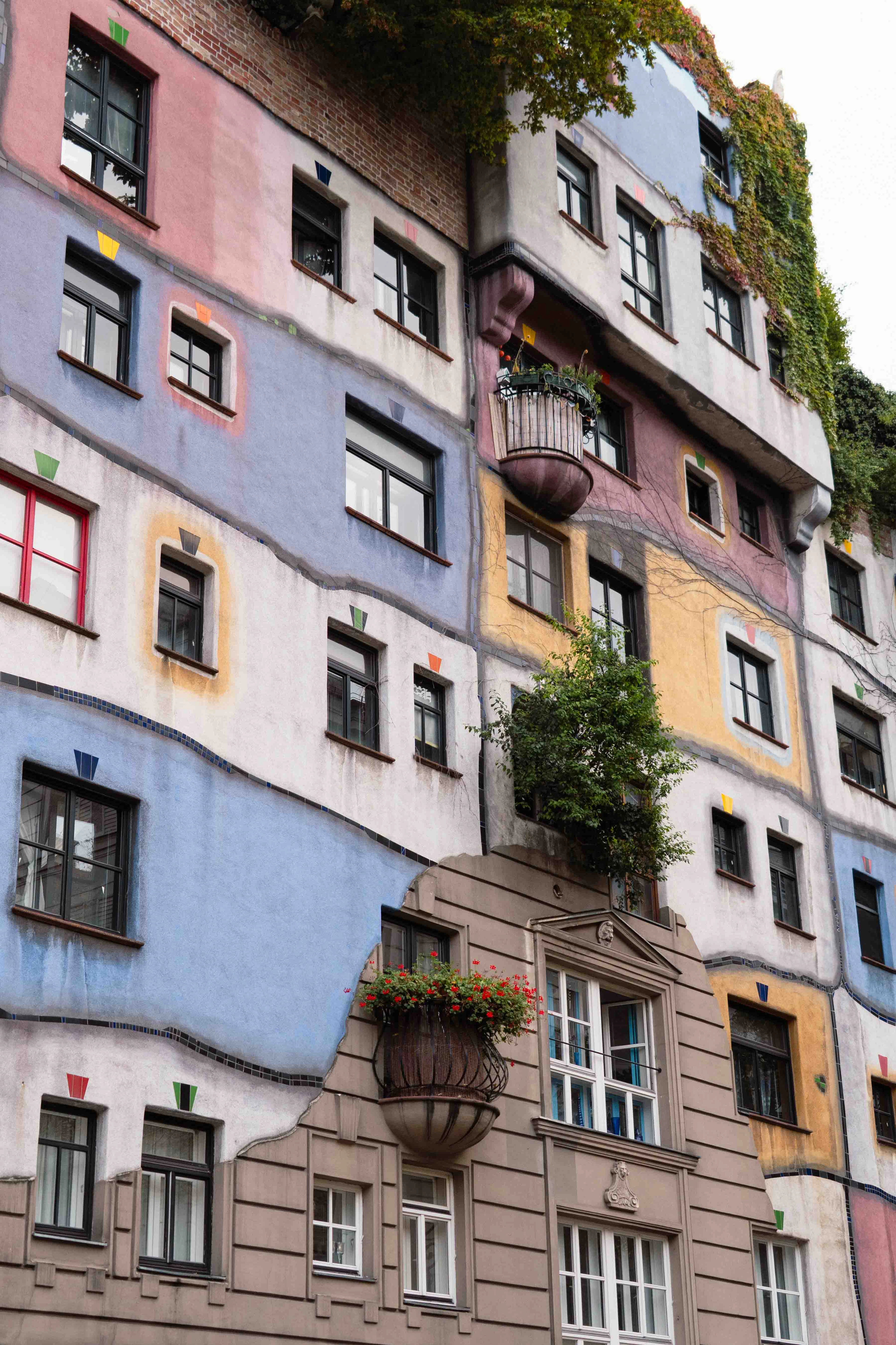 The Hundertwasserhaus in Vienna seen close up, its facade a patchwork of pink, blue, white and ochre panels divided by wavy borders, windows of different sizes in mismatched frames, a bulbous balcony spilling red geraniums and small trees growing straight out of the building.