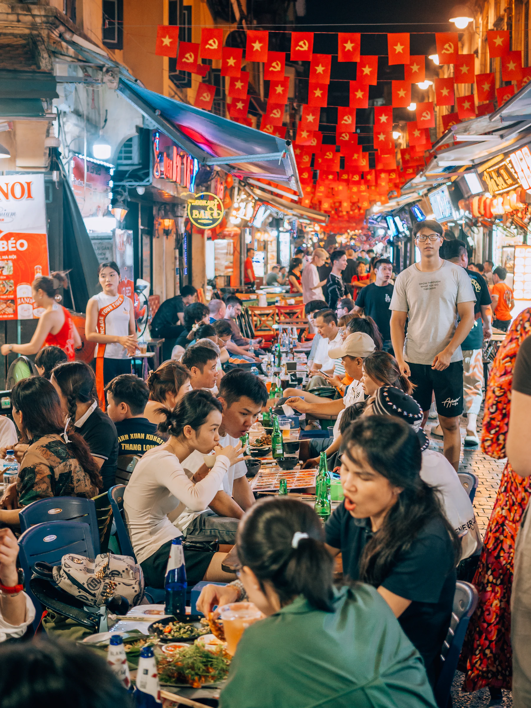 A packed street-food market at night, diners eating at low tables under strings of red flags