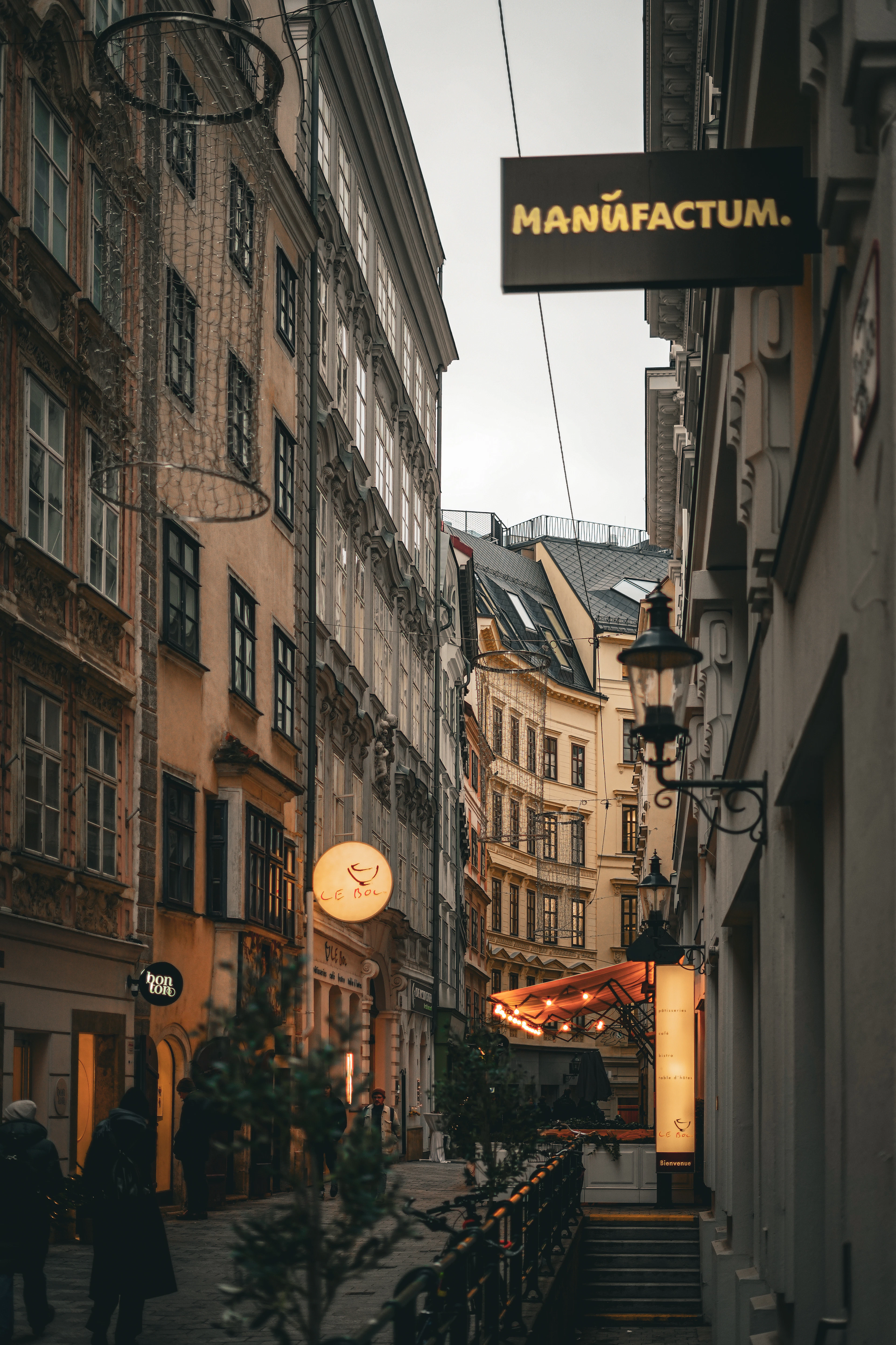 A narrow Vienna backstreet in flat winter light, tall old houses leaning in on both sides with hanging shop signs and an ornate wrought-iron street lamp, warm lit shop windows below and an awning strung with bulbs over a terrace at the far end.