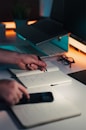 Hands writing in notebook near laptop and phone.