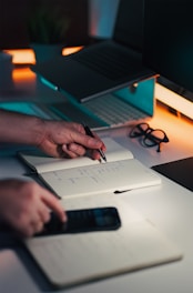 Hands writing in notebook near laptop and phone.