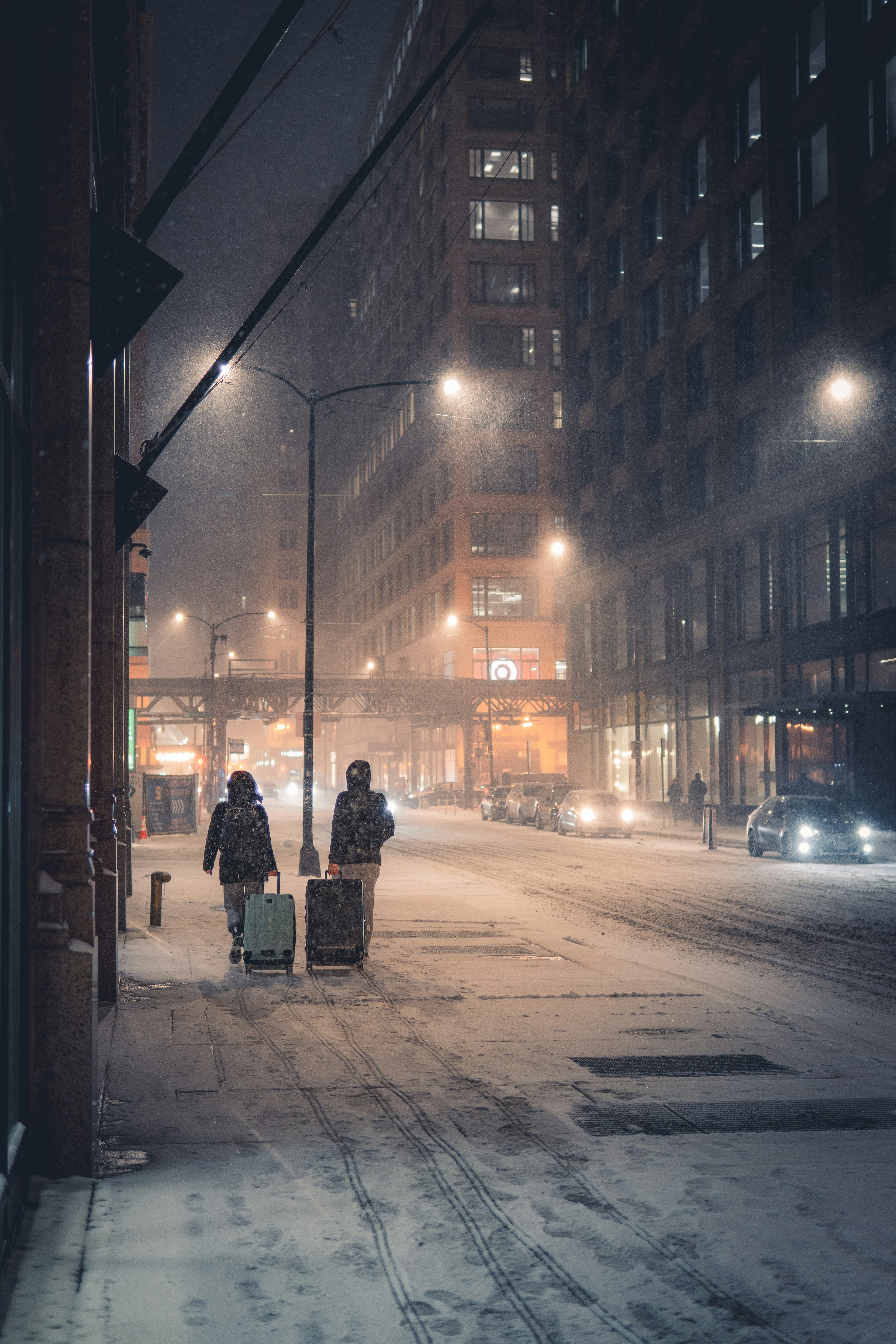 Two people with luggage walk down snowy city street