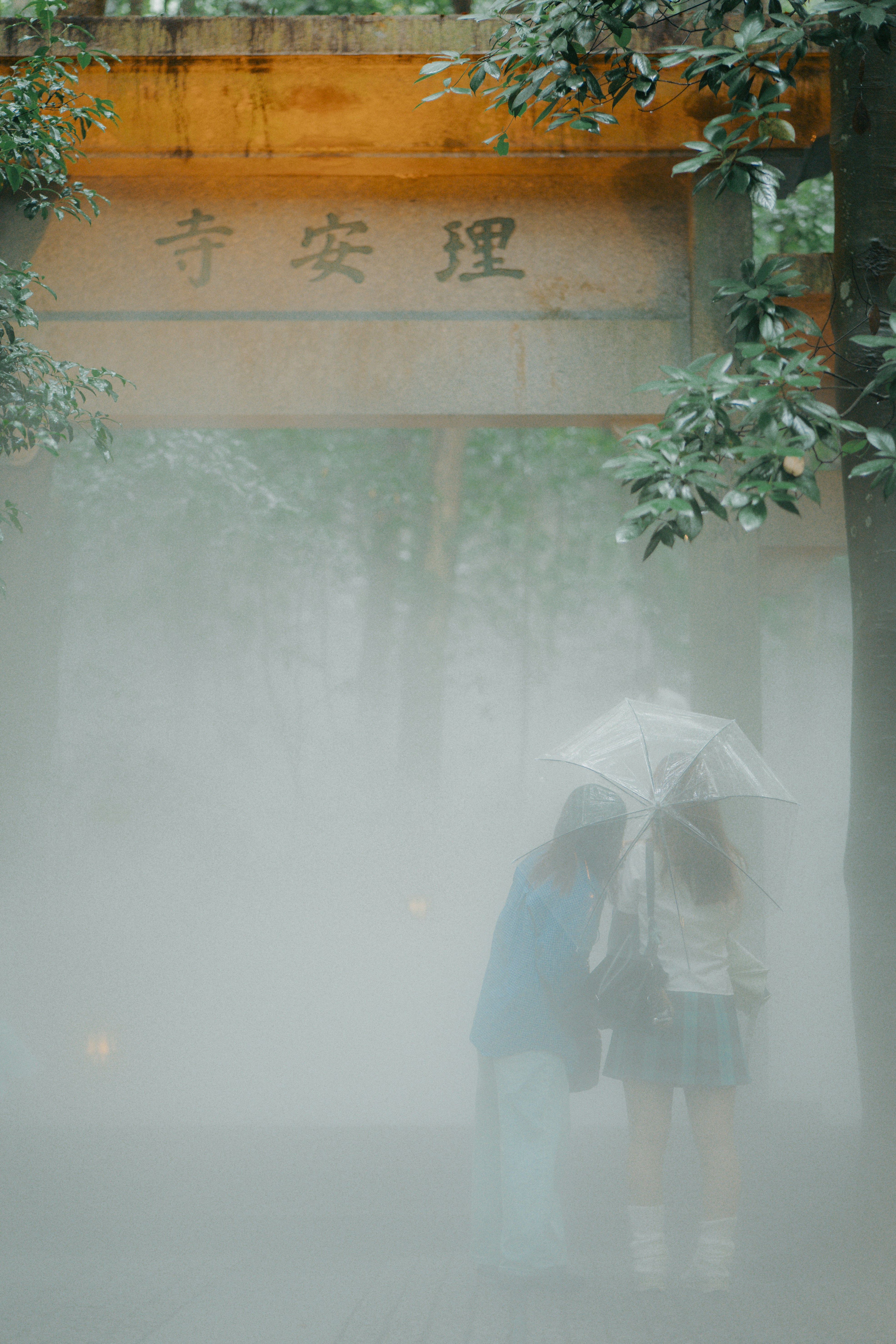 Two people with umbrellas walk through misty temple entrance