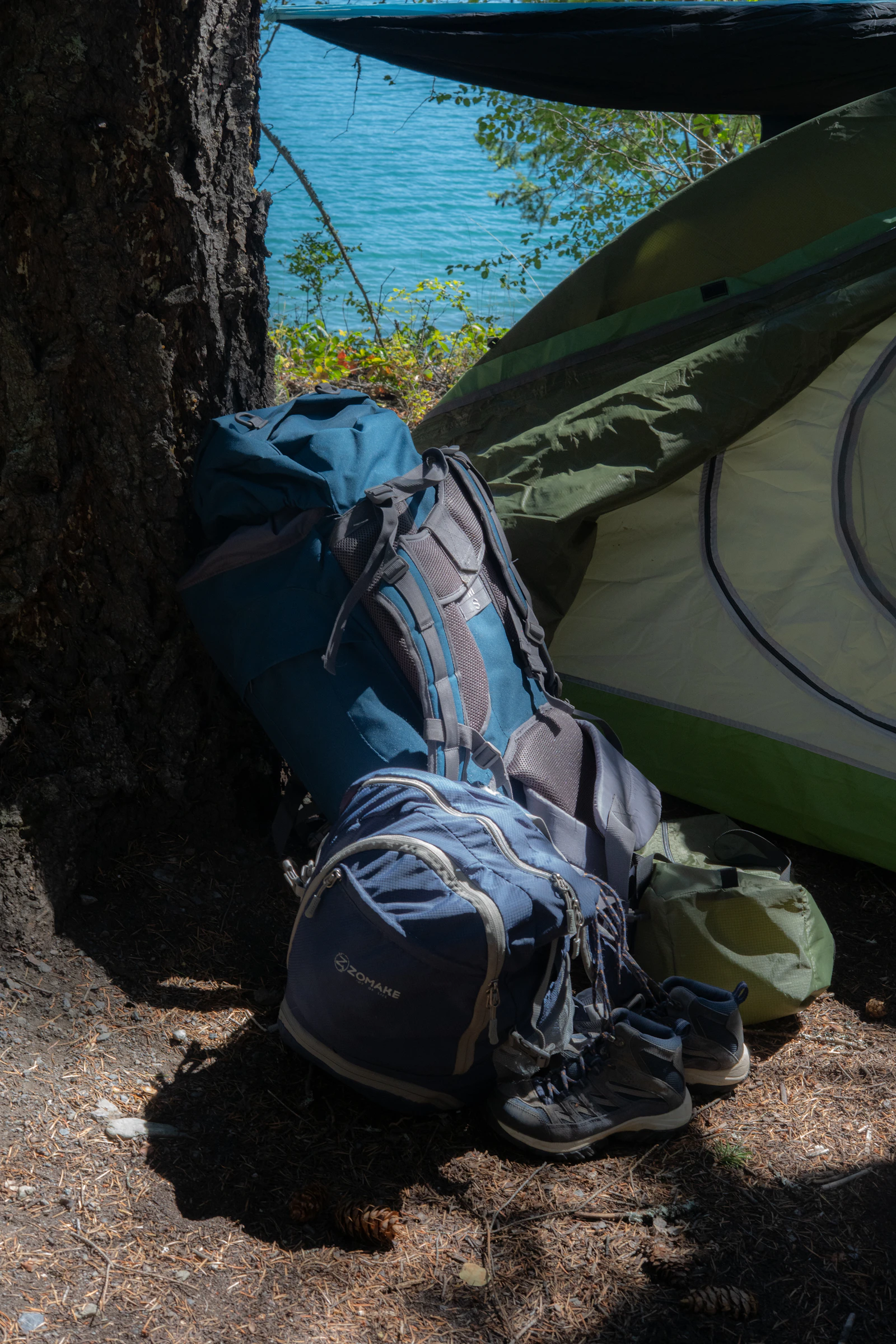 Camping backpack, footwear, and tent arranged beside a lake