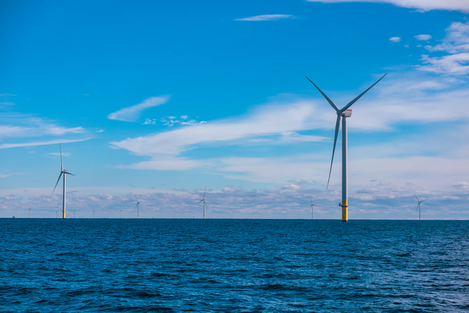 Six wind turbines stand tall on calm ocean waters with white-gray turbines against blue sky