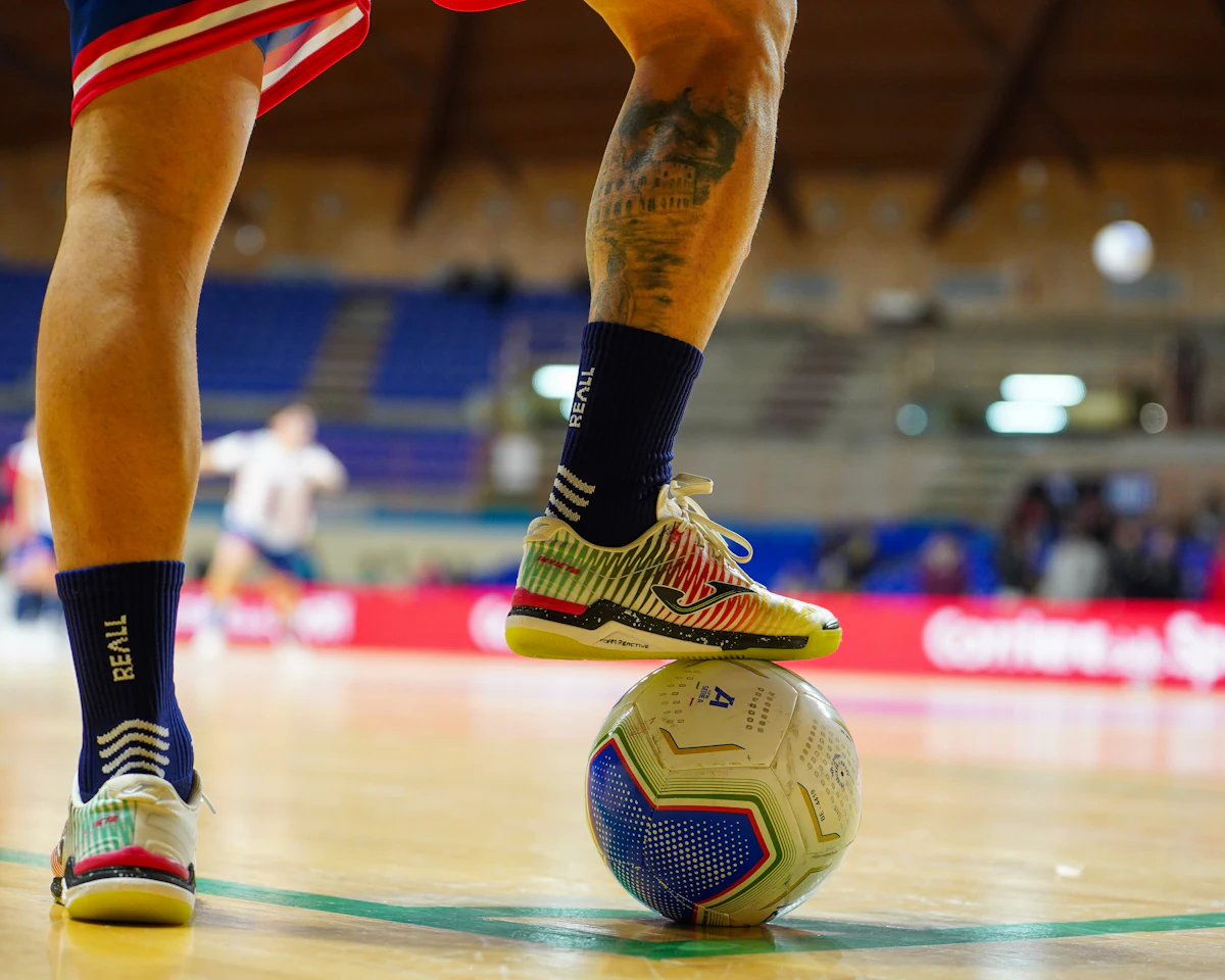 Pemain futsal menguasai bola di lapangan gor kayu, lampu sorot dari langit-langit ruangan tertutup