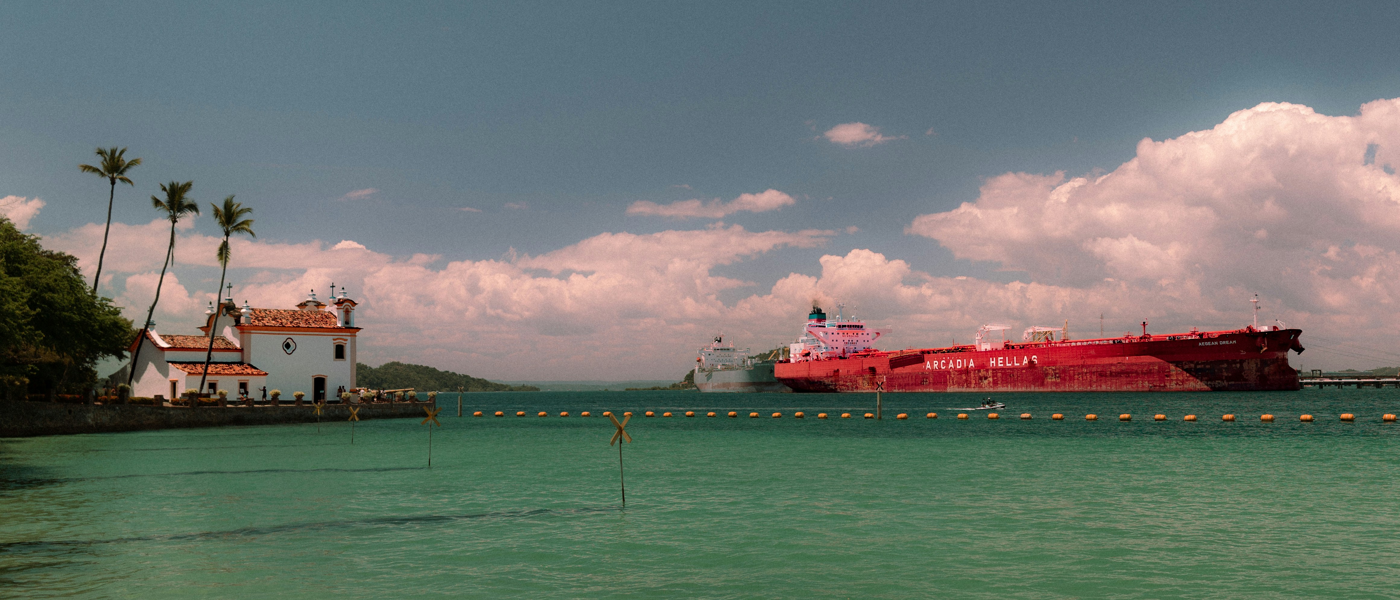 A large red ship sails on turquoise water near shore.