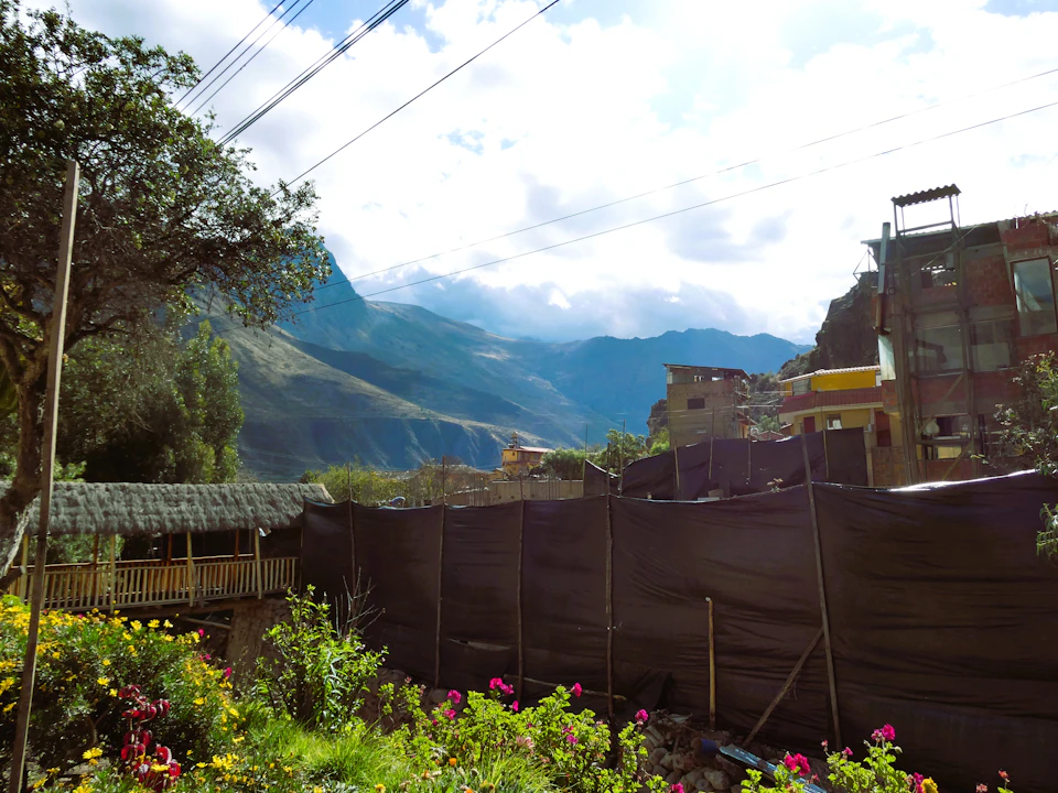 The image depicts a serene mountainous landscape with a lush green valley and a clear blue sky dotted with fluffy white clouds. The mountains, covered in trees and shrubs, stretch across the horizon, creating a sense of depth and vastness. In the foreground, there is a black fence enclosing a small garden filled with vibrant flowers, adding a touch of color to the scene. On the left side of the image, a wooden bridge can be seen, possibly leading to another part of the valley or beyond.