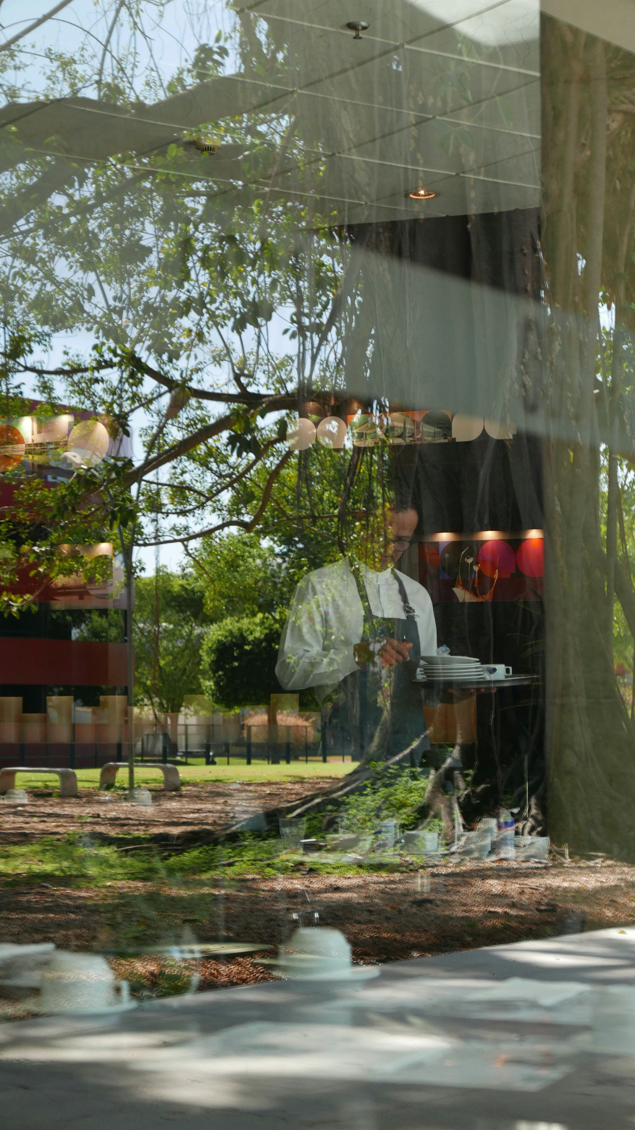 Koch in Schürze hält Teller in einem Caféfenster und spiegelt sich.