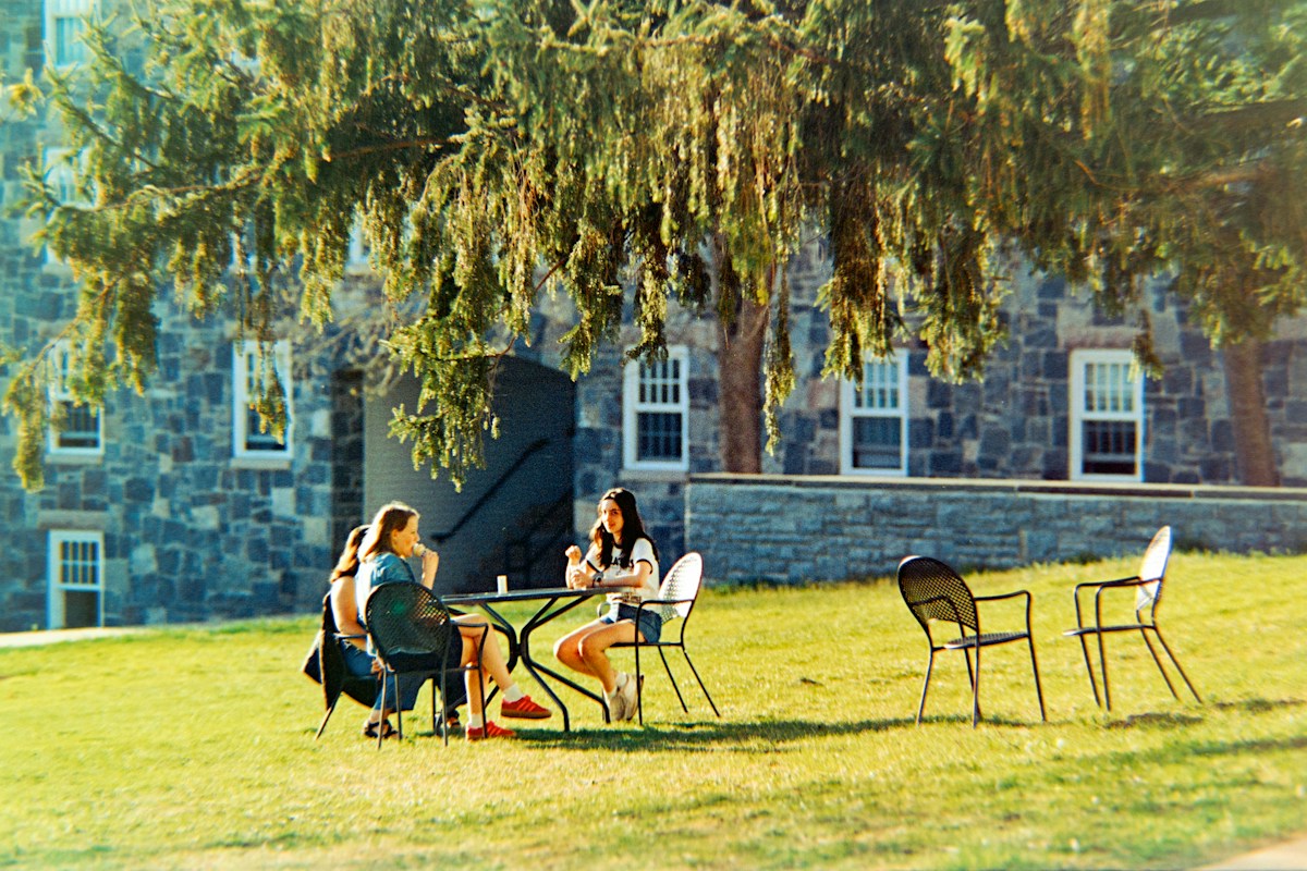 Two women sit at a table outside a stone building, discussing college club ideas and planning their first event.
