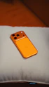 An orange smartphone rests on a light-colored pillow.