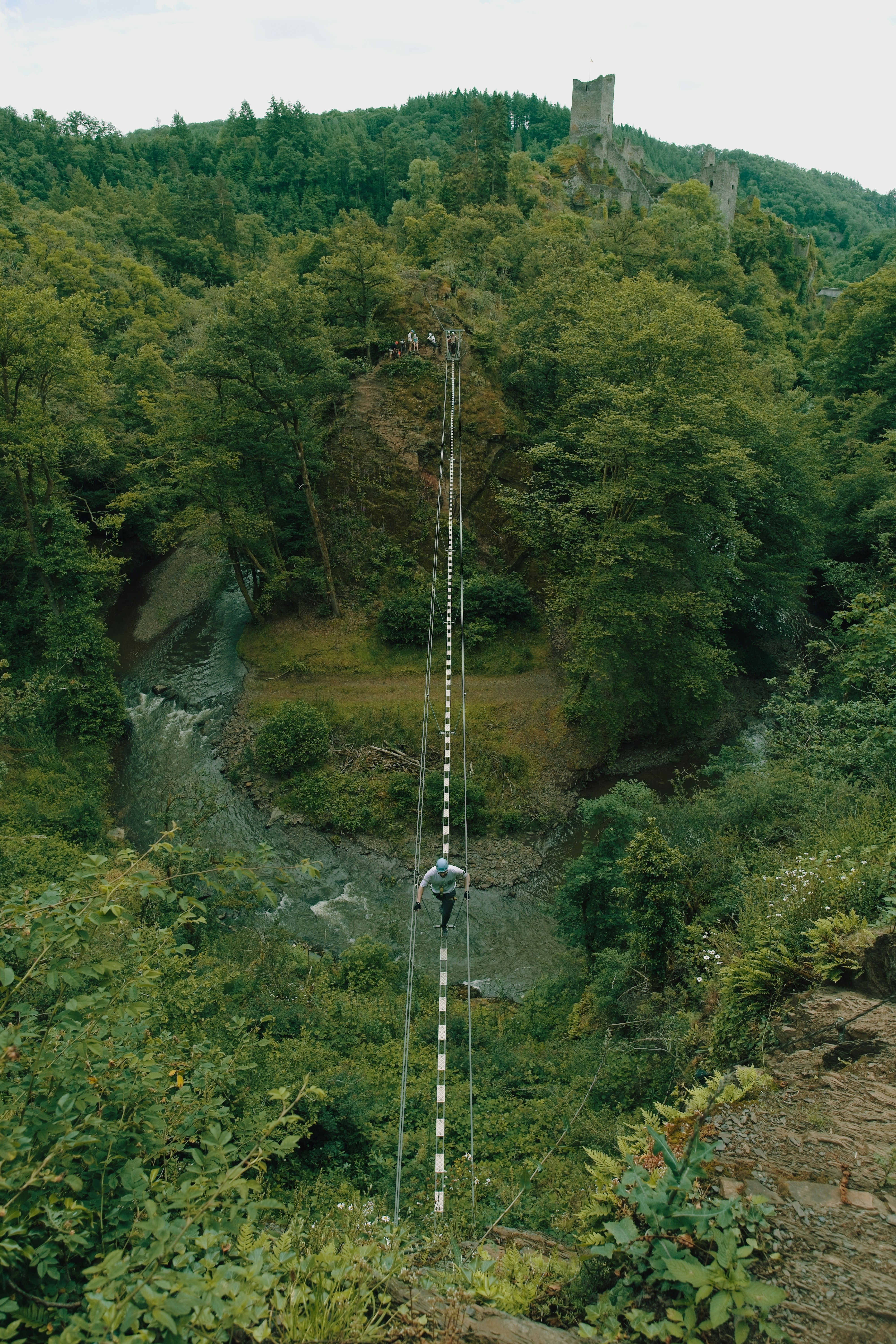 Person rappelling down a steep forested gorge towards ruins.