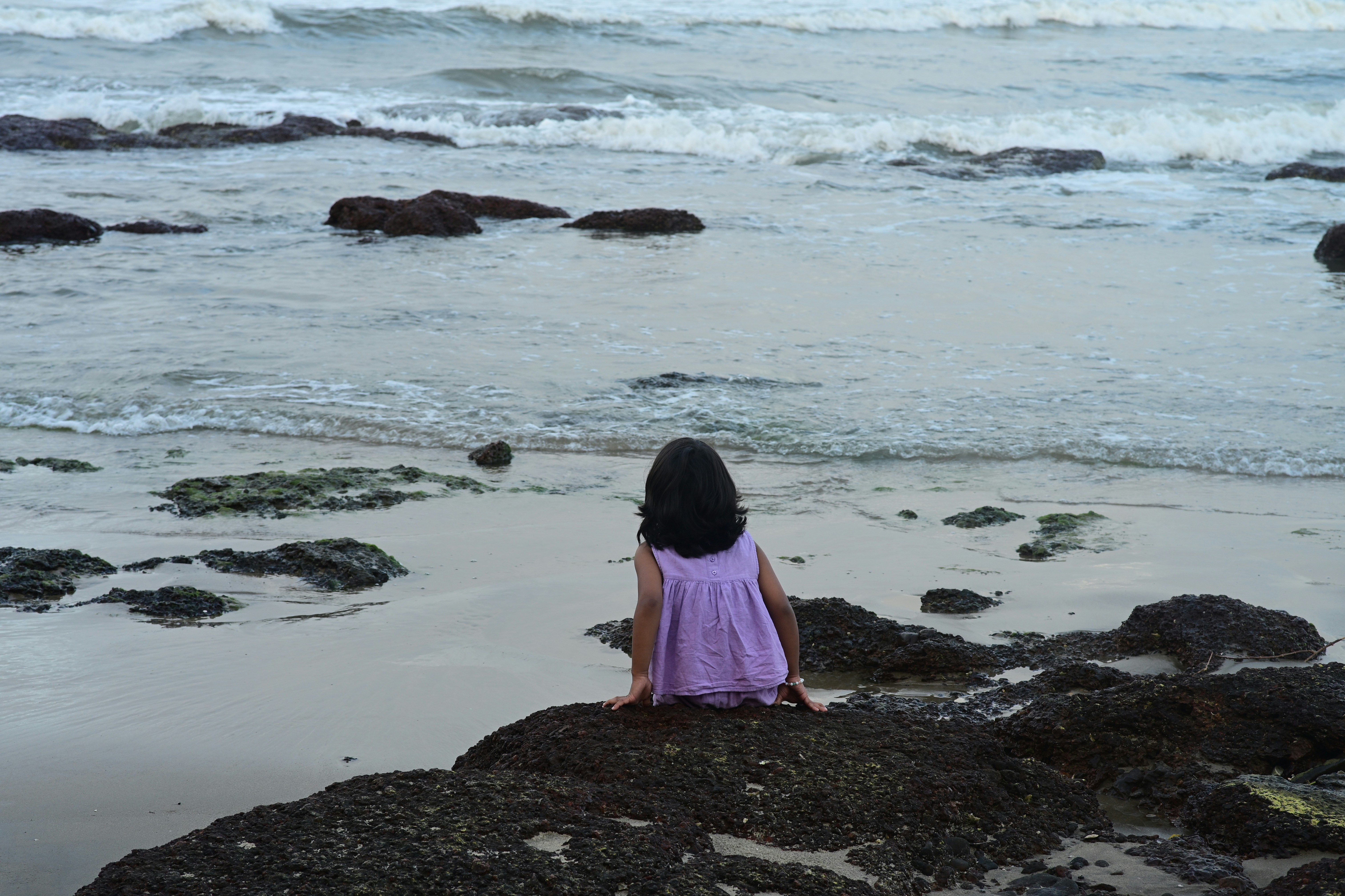 Girl in purple dress sits on rocks by the ocean.