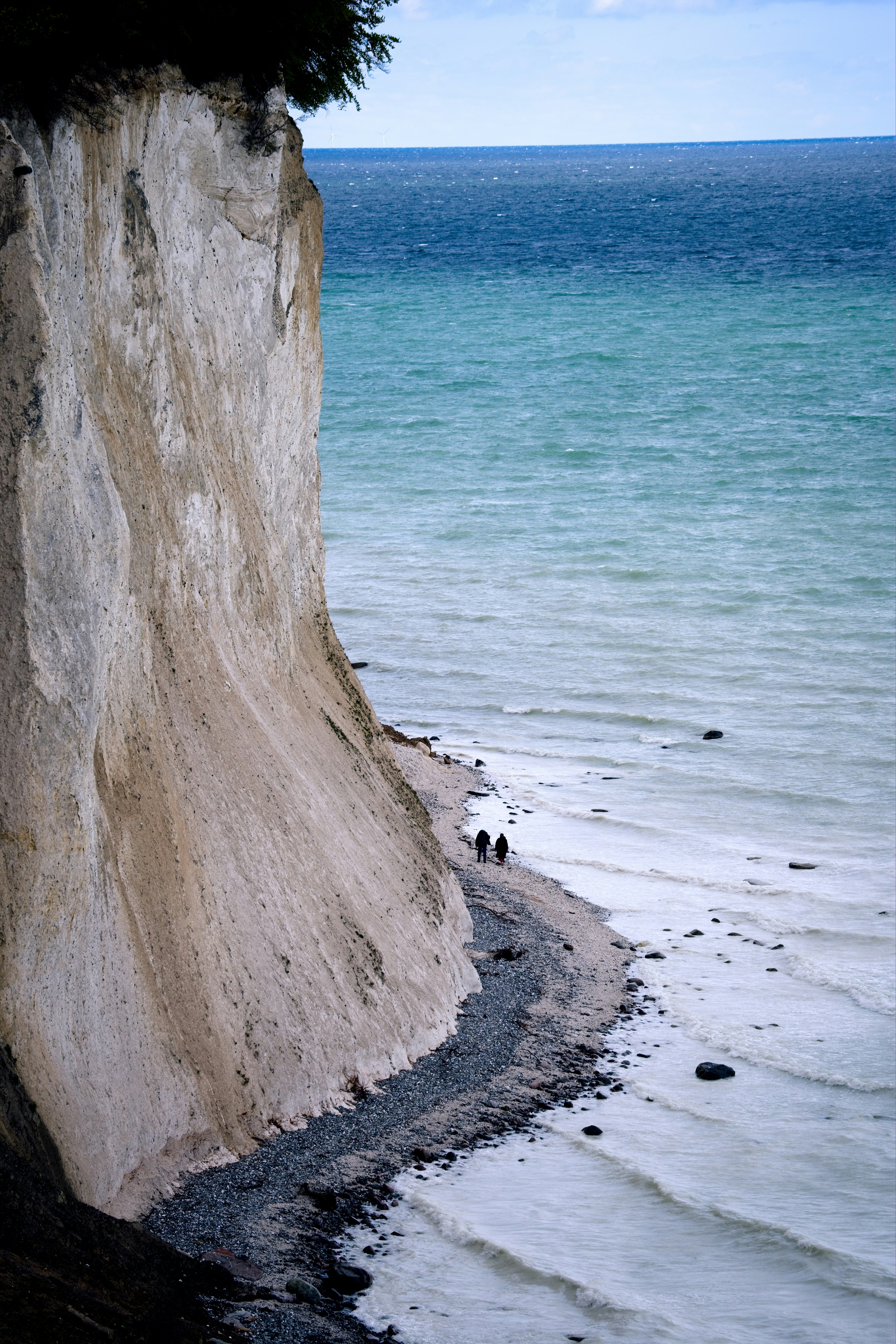 Two people walk along a rocky beach near cliffs.