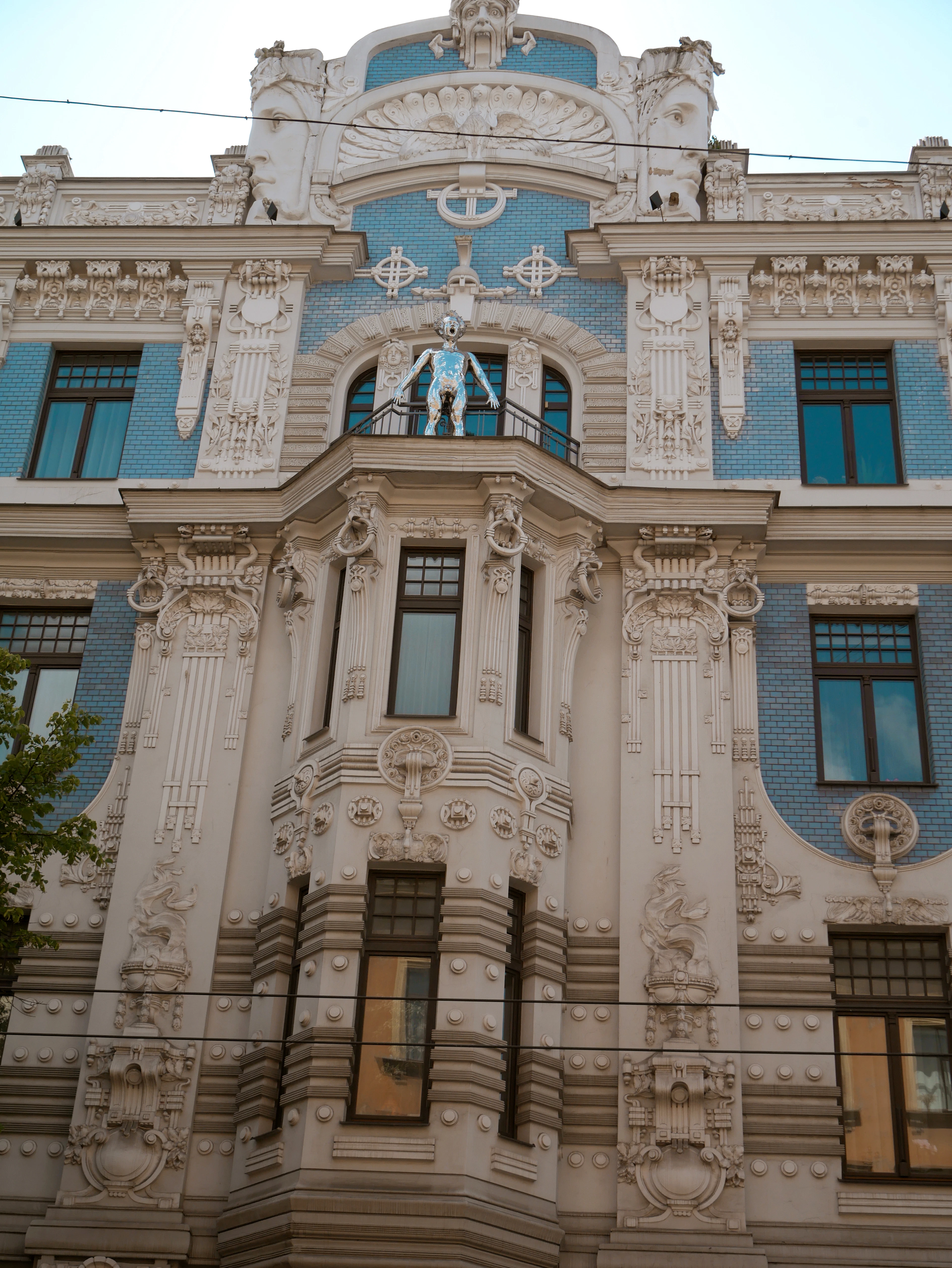 The ornate Art Nouveau facade of an Alberta iela apartment house in Riga, its cream stucco crowded with sculpted masks, wreaths and slender pilasters against panels of blue glazed brick, a figure standing on the small balcony beneath the gable.