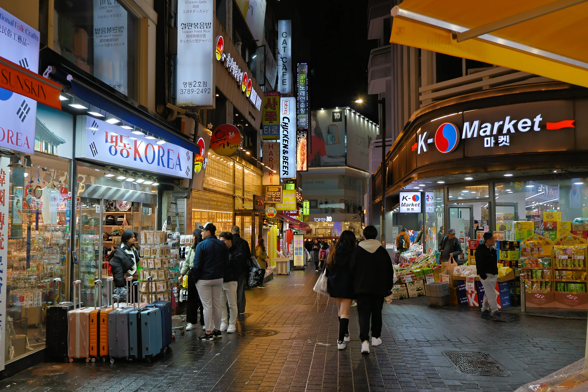 People walk along a brightly lit Myeongdong shopping street lined with Korean shops and Market grocery storefronts in the evening.