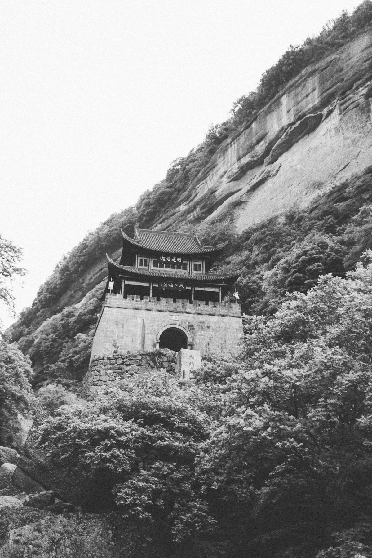 The gate tower of Jianmen Pass below a sheer rock cliff, in black and white, with trees in front