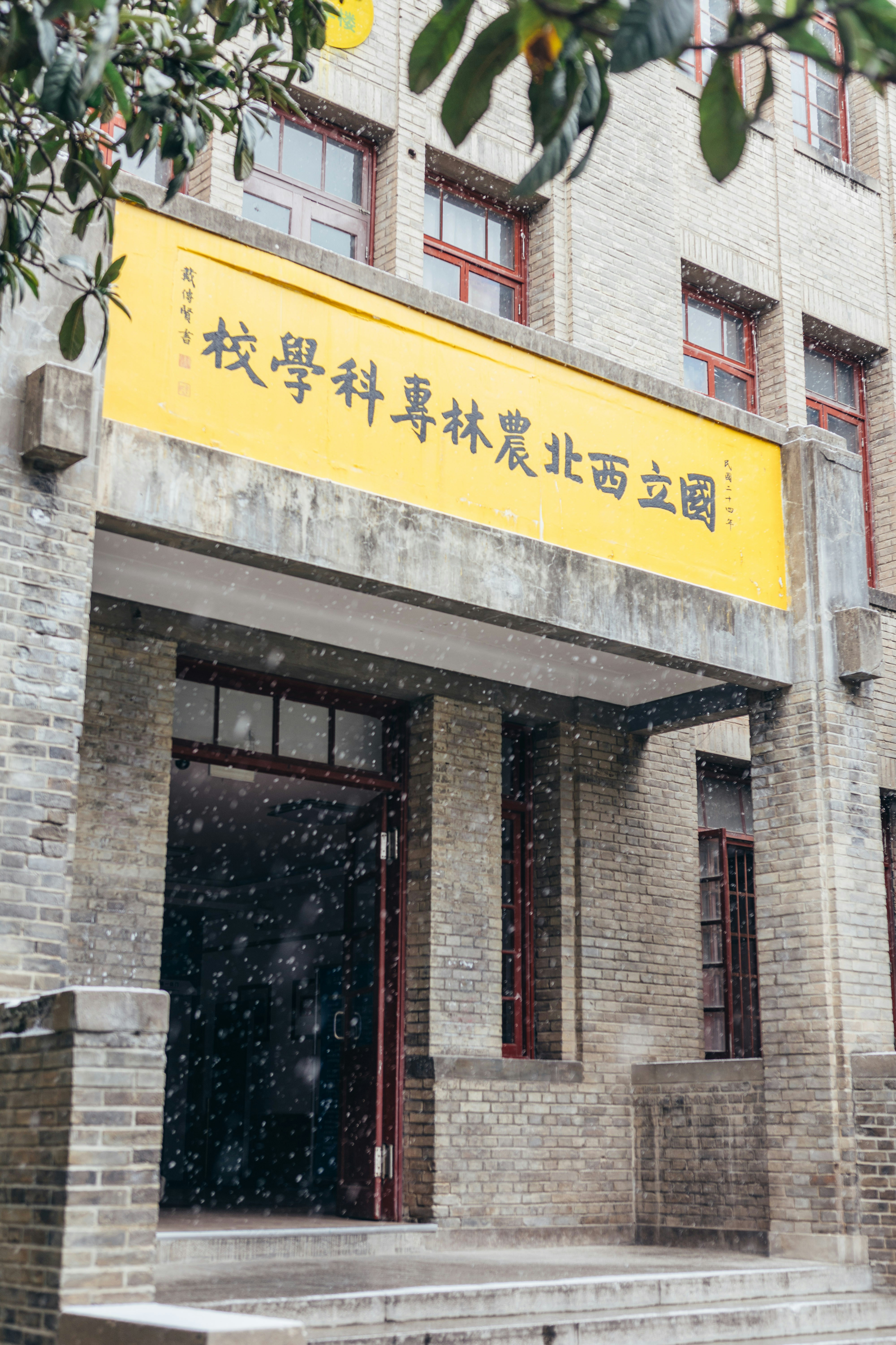 Snow falls on a brick building entrance with yellow signage.