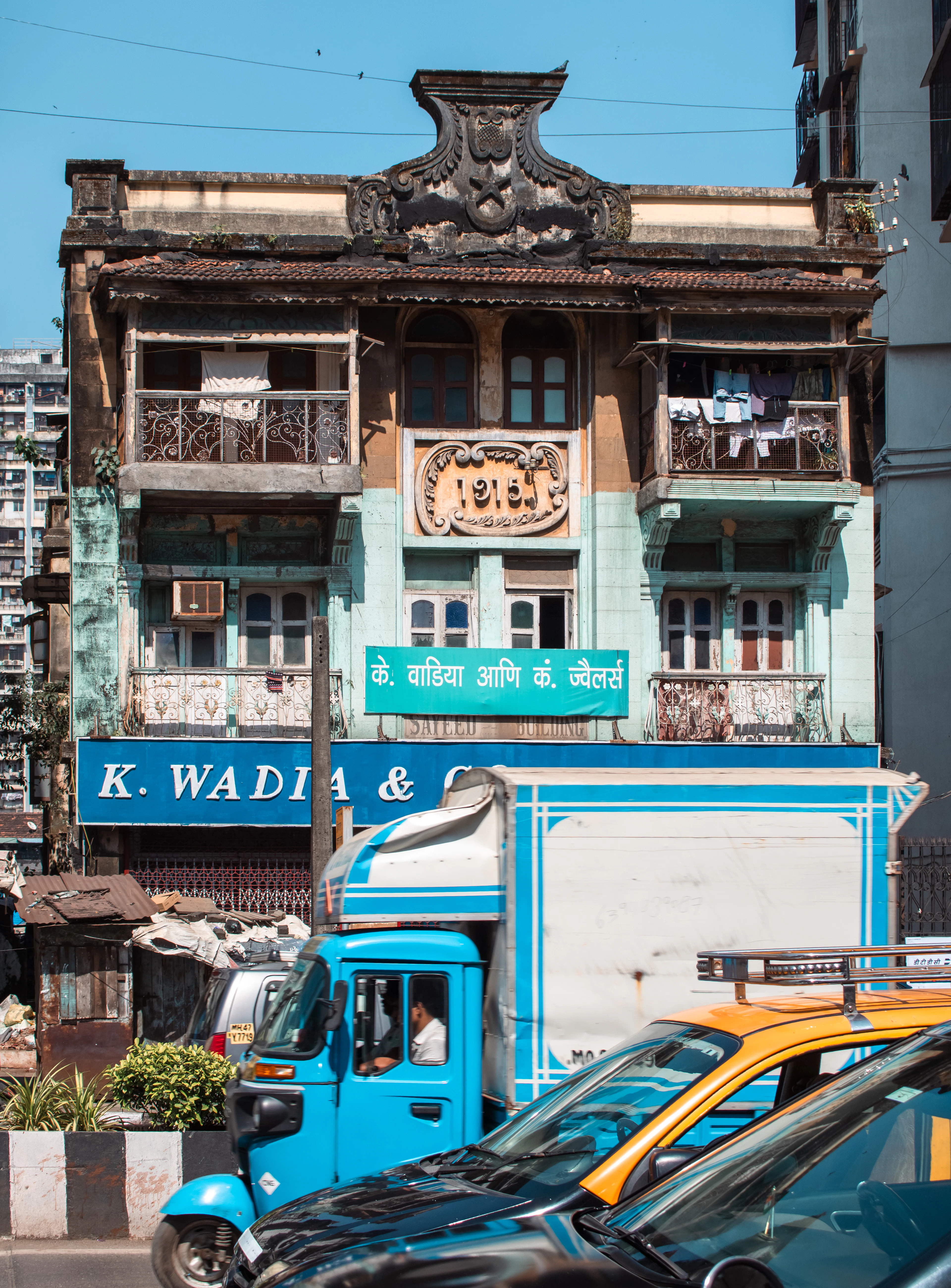 A weathered colonial-era building with balconies and hanging laundry stands above a shop, with an auto-rickshaw and taxis below.