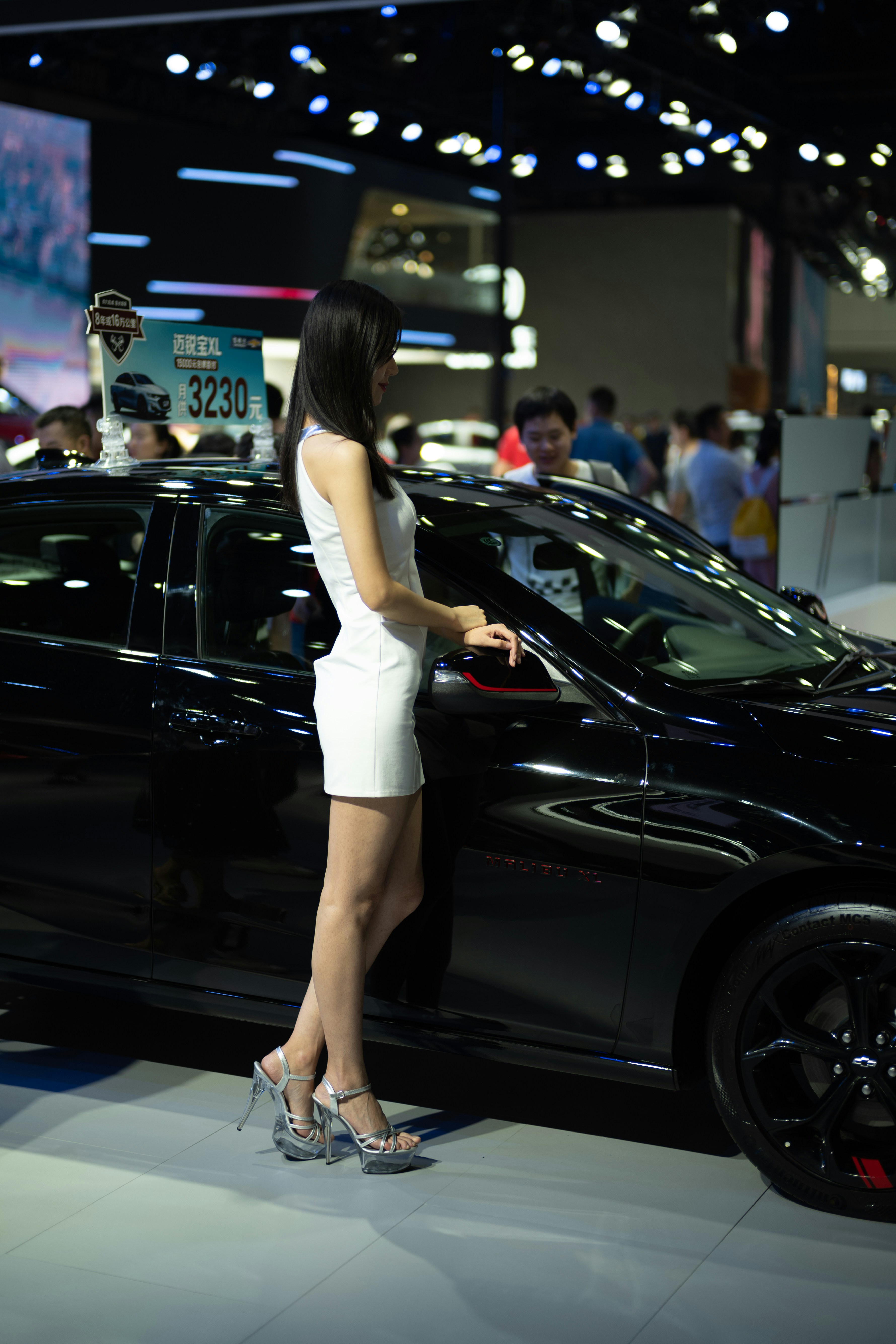 Woman in white dress posing by black car