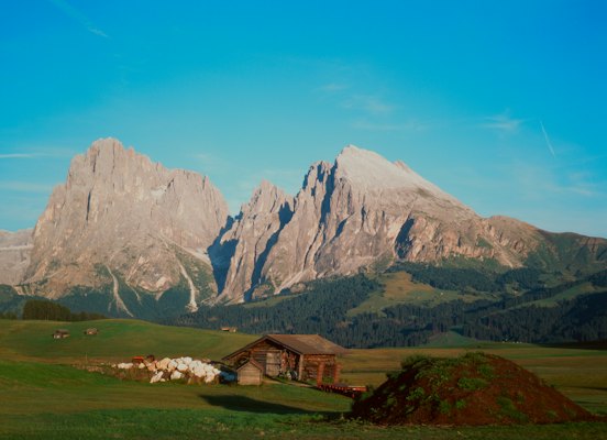 A rustic cabin sits in a green meadow below mountains — Dolomites, Italy