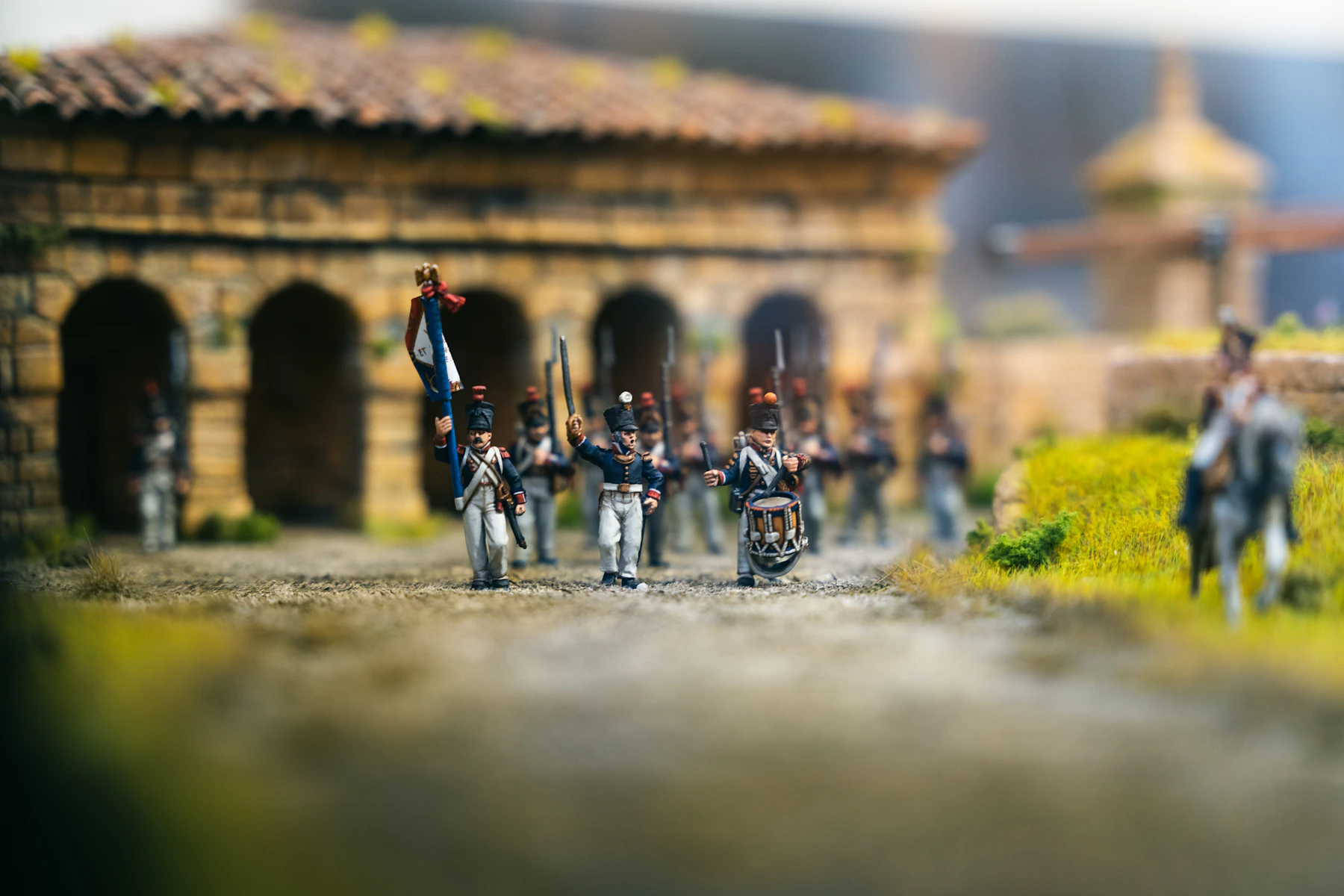 Painted model soldiers in blue coats marching with a flag past a stone building on a scenic base