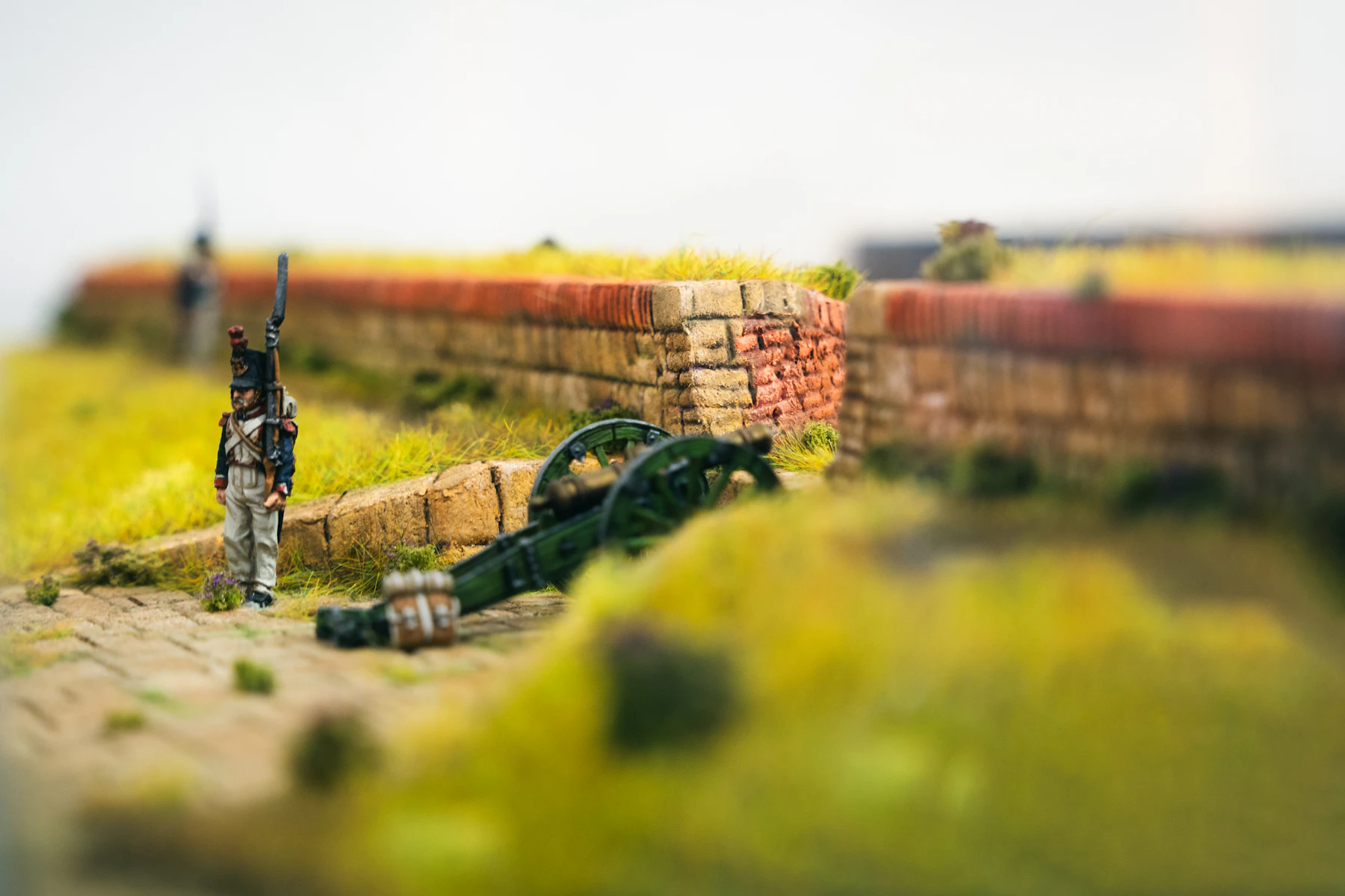 A miniature soldier and a green field cannon beside a low red brick wall, with out-of-focus yellow-green grass in the foreground