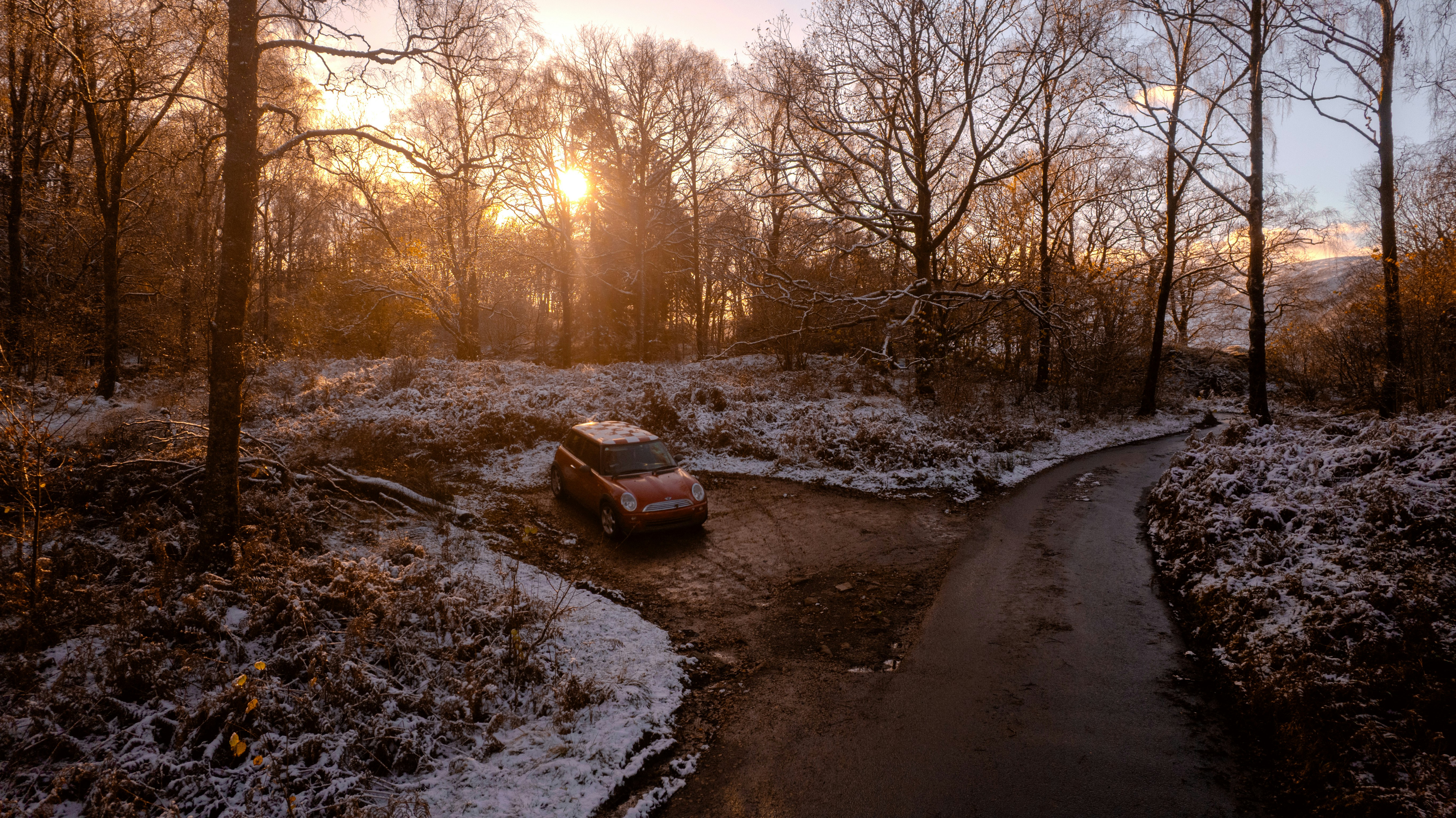 A car drives through a snowy forest at sunrise.
