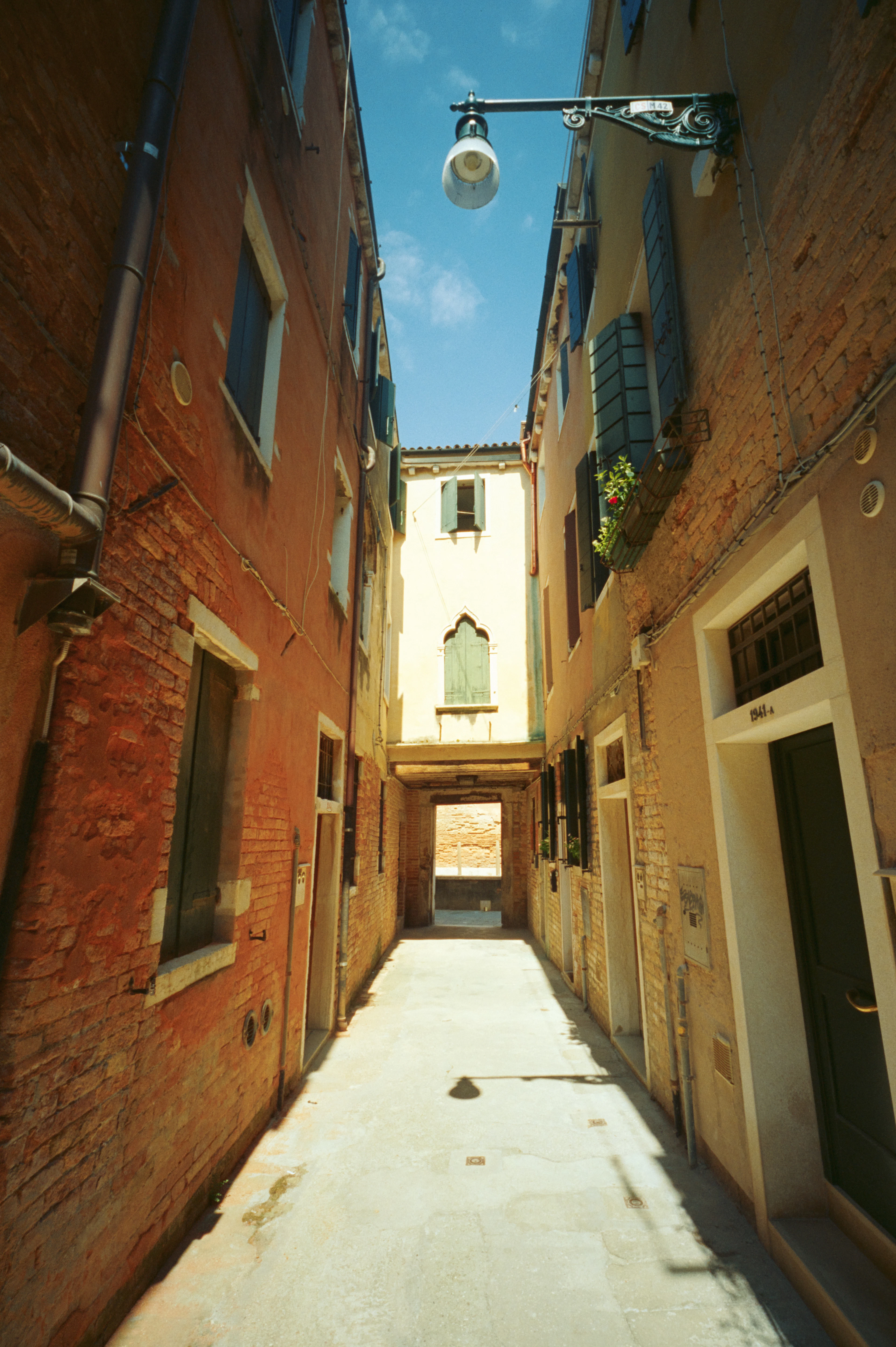 A narrow Venetian calle in Cannaregio at midday, sun striking worn brick and ochre plaster walls, an iron street lamp overhead and a shaded sottoportego at the far end