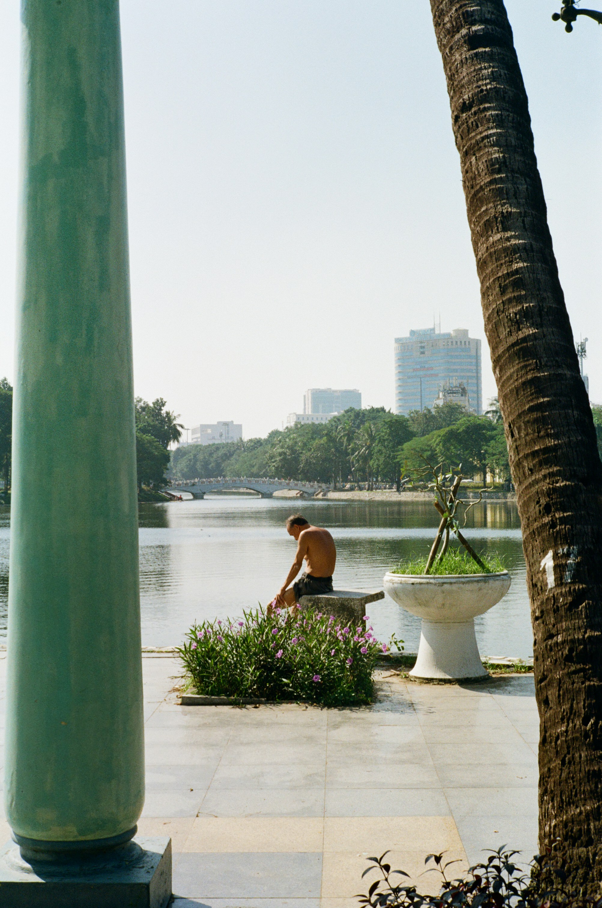 Man sitting by a calm lake with buildings in background.
