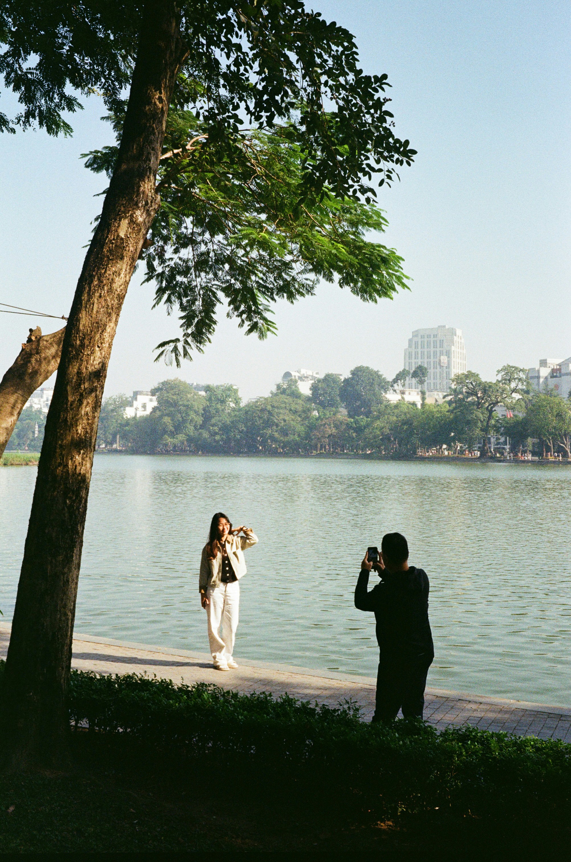 Man photographs woman by a lake with trees.