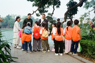 Adults guide children wearing orange vests by a lake.