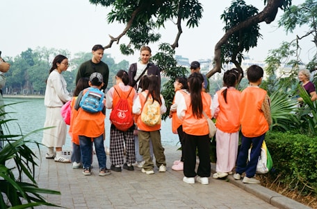 Adults guide children wearing orange vests by a lake.