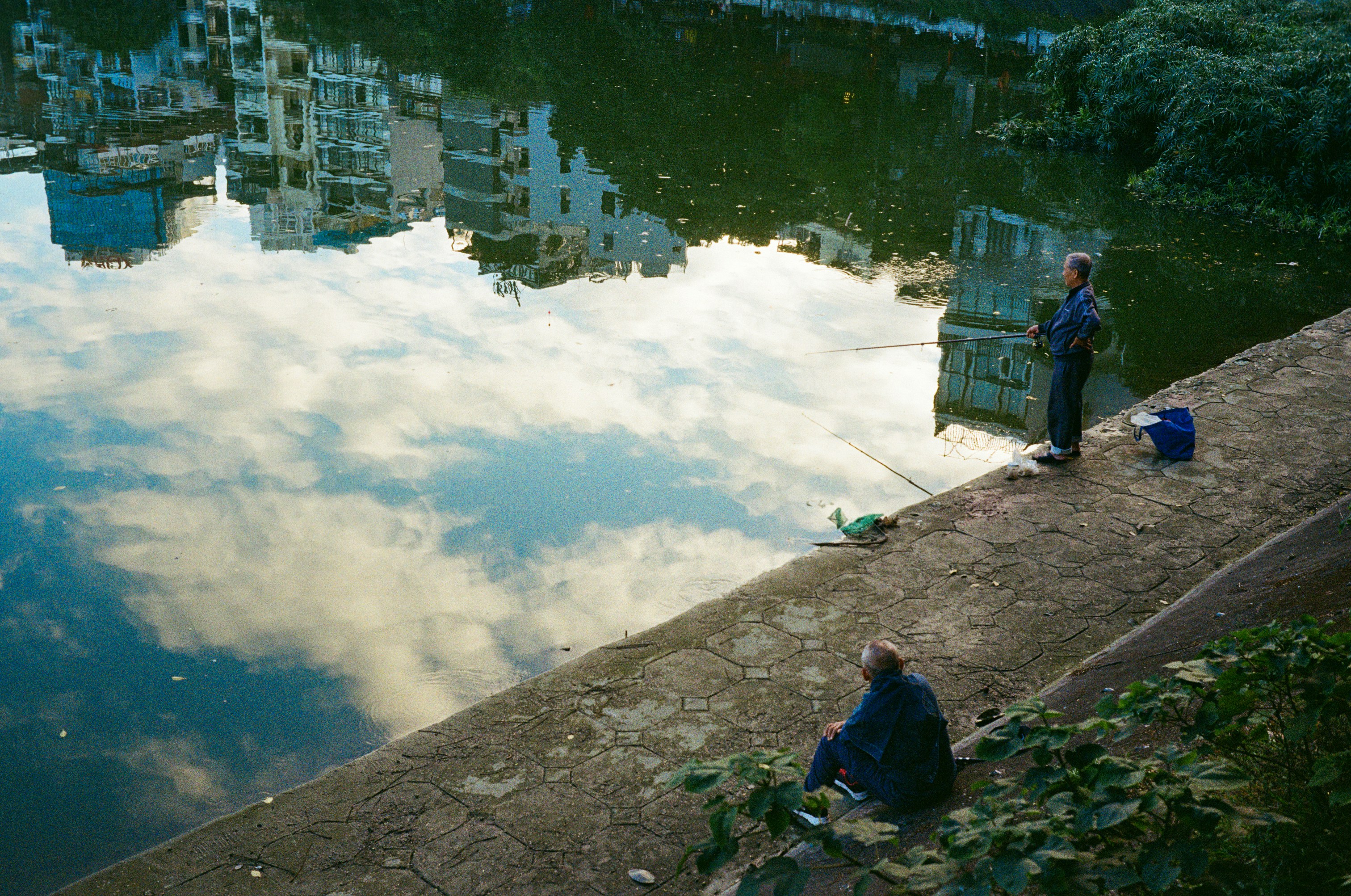 Two people fishing by a reflective lake with buildings.