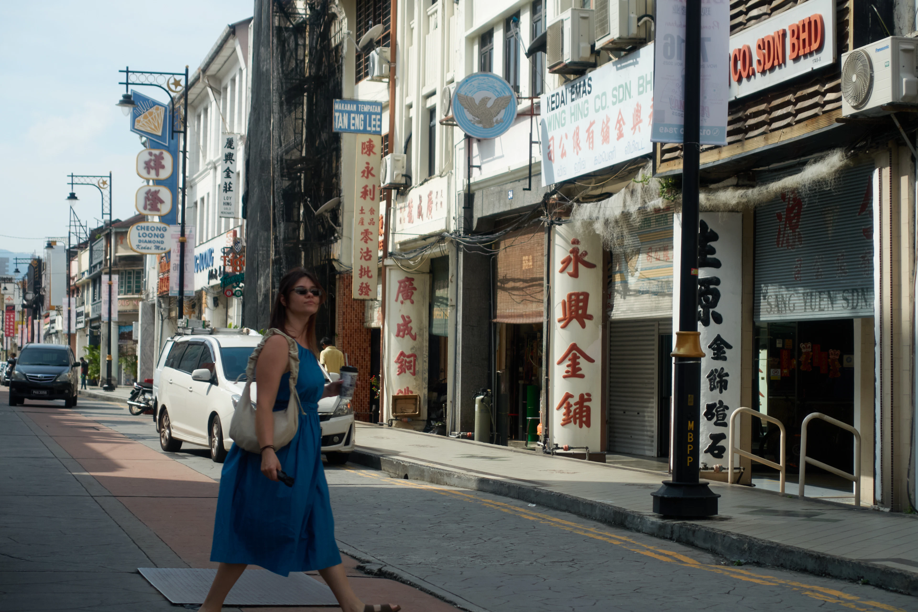A woman in a blue dress walks past a row of old shophouses covered in Chinese-language signboards.