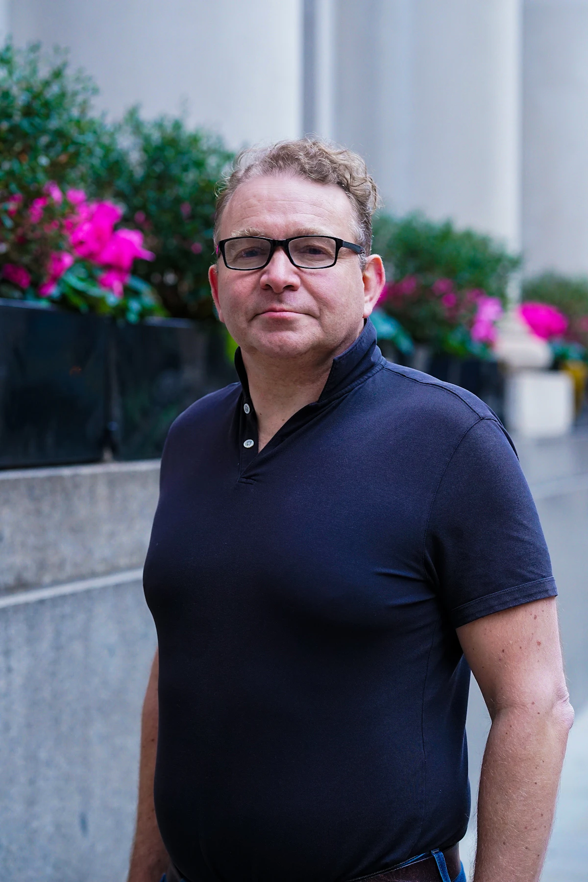 Homem de óculos com camisa polo azul-marinho lisa, sem logo aparente, parado numa calçada com flores ao fundo