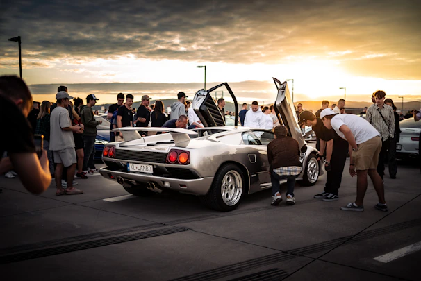 People gathered around a silver sports car at sunset.