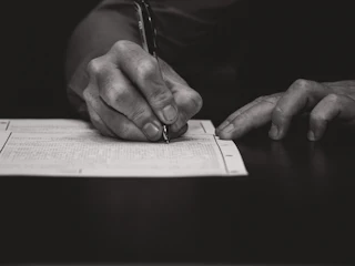 Black-and-white close-up of an older adult's hands holding a pen over a dense printed ledger page
