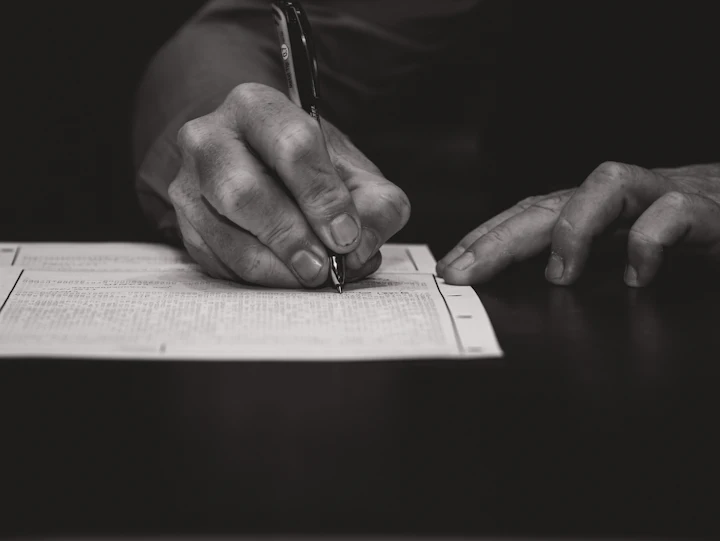 Black-and-white close-up of an older adult's hands holding a pen over a dense printed ledger page